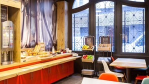Un café avec un comptoir en bois, une machine à café et une vue sur l’extérieur à travers une fenêtre ornée de barreaux en fer forgé.