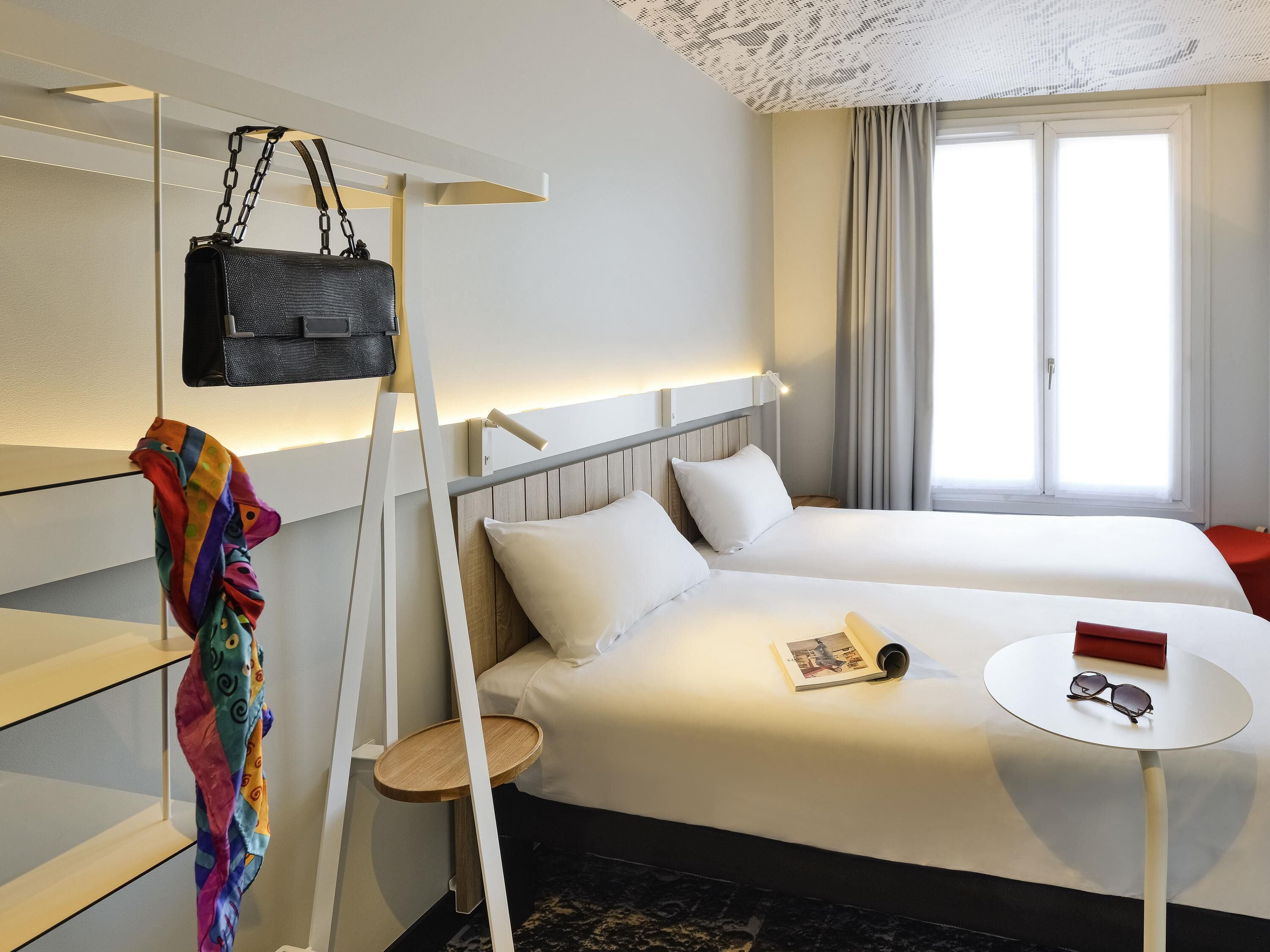 Una habitación de hotel con una cama, una mesita de noche con vasos, un bolso colgado y una bufanda colorida.