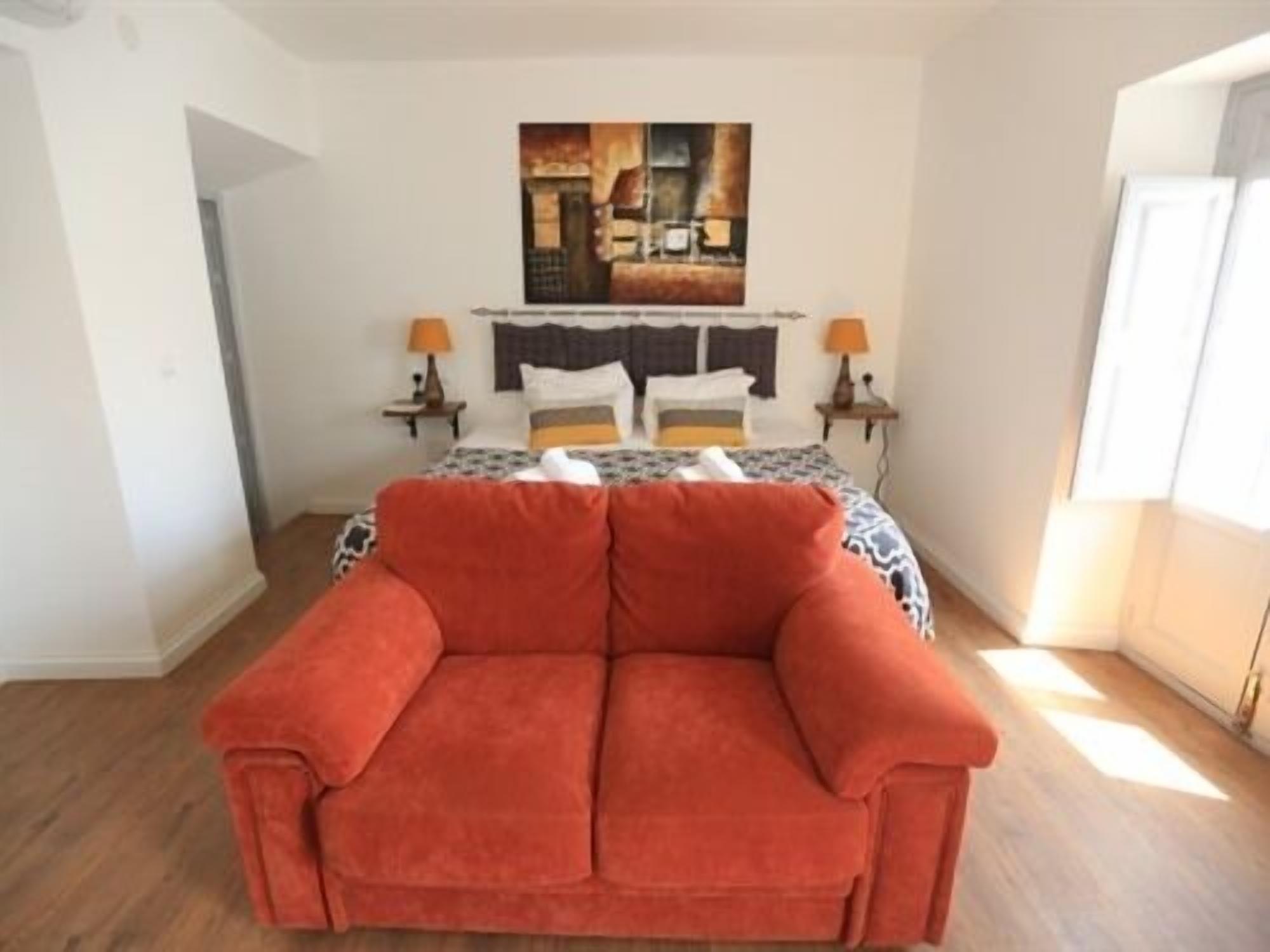 Quarto com cama, sofá vermelho, duas mesas de cabeceira com abajures e um quadro na parede.