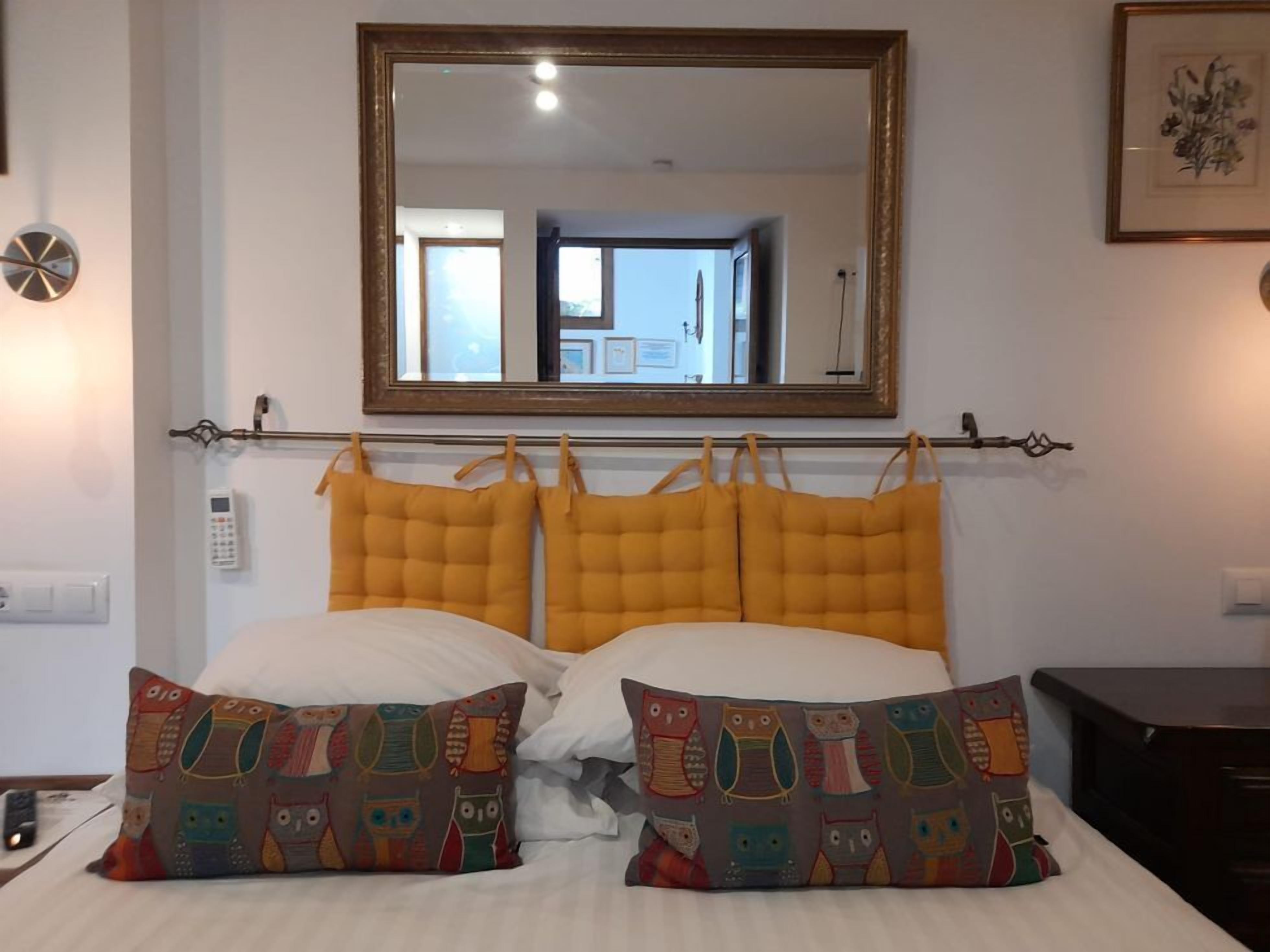 A hotel room with a bed, two matching pillows, a bedside table, a bedside lamp, a wall-mounted telephone, a wall clock, and a large mirror above the bed.