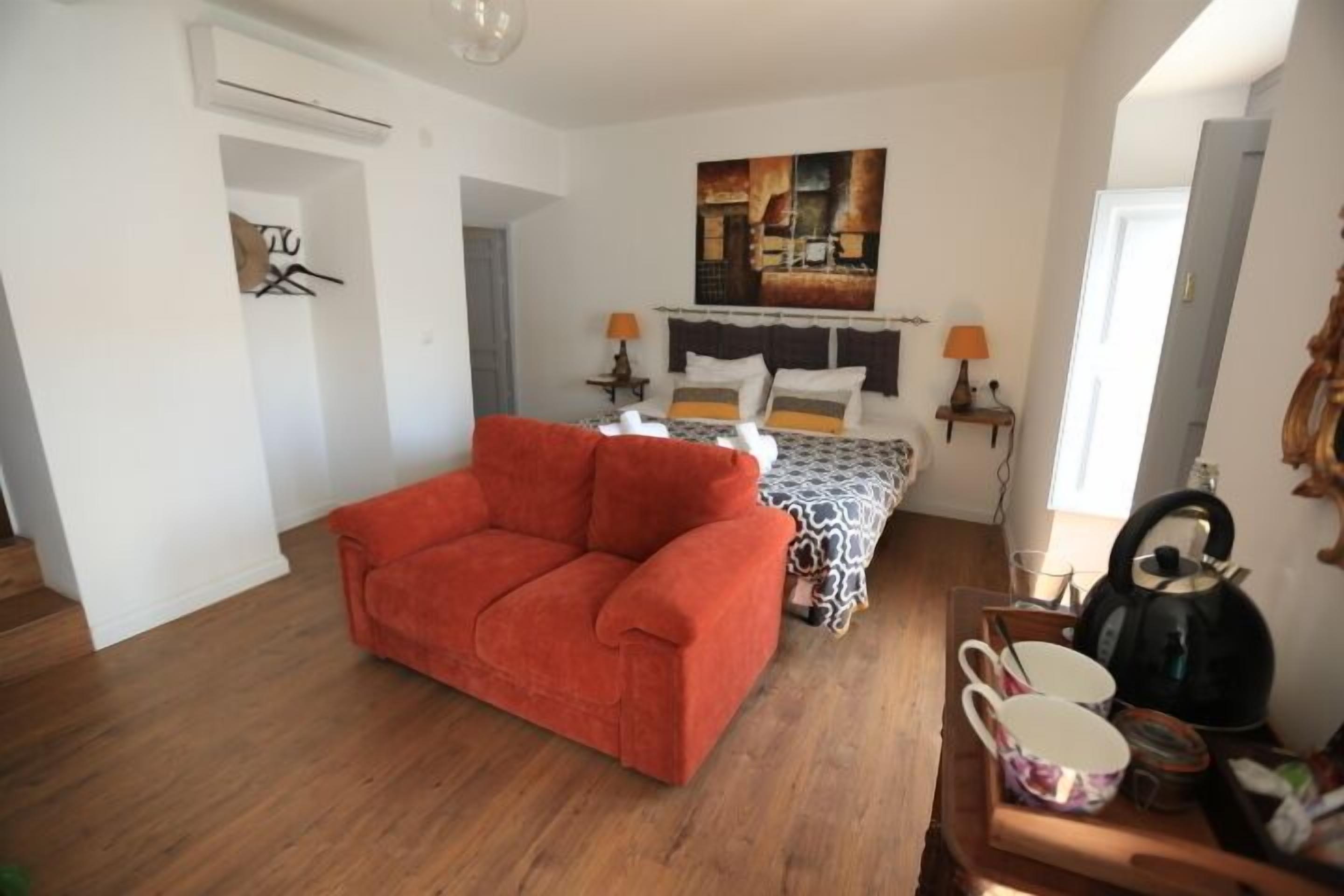 A bedroom with a bed, a red sofa, a bedside table with a kettle, and a painting on the wall.