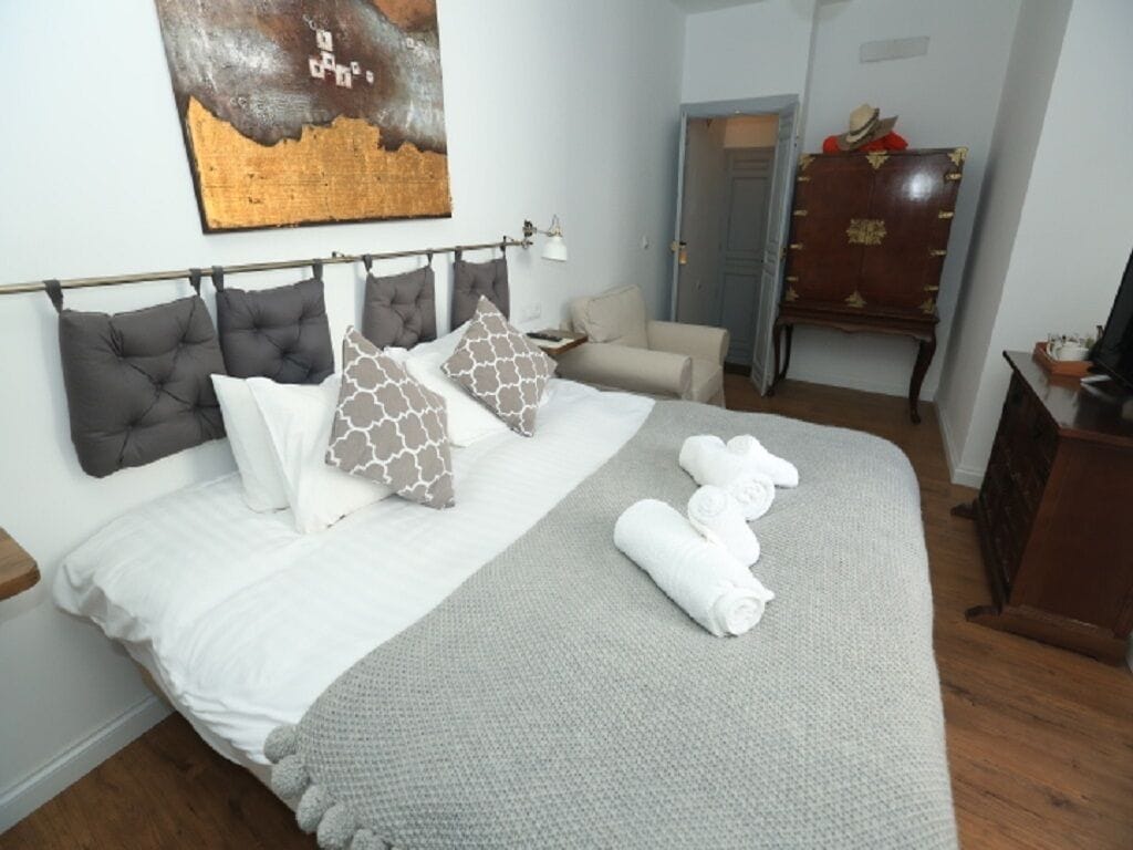 A neatly made bed with a grey blanket and rolled towels, a headboard with multiple pillows, a wooden dresser, and a painting on the wall.