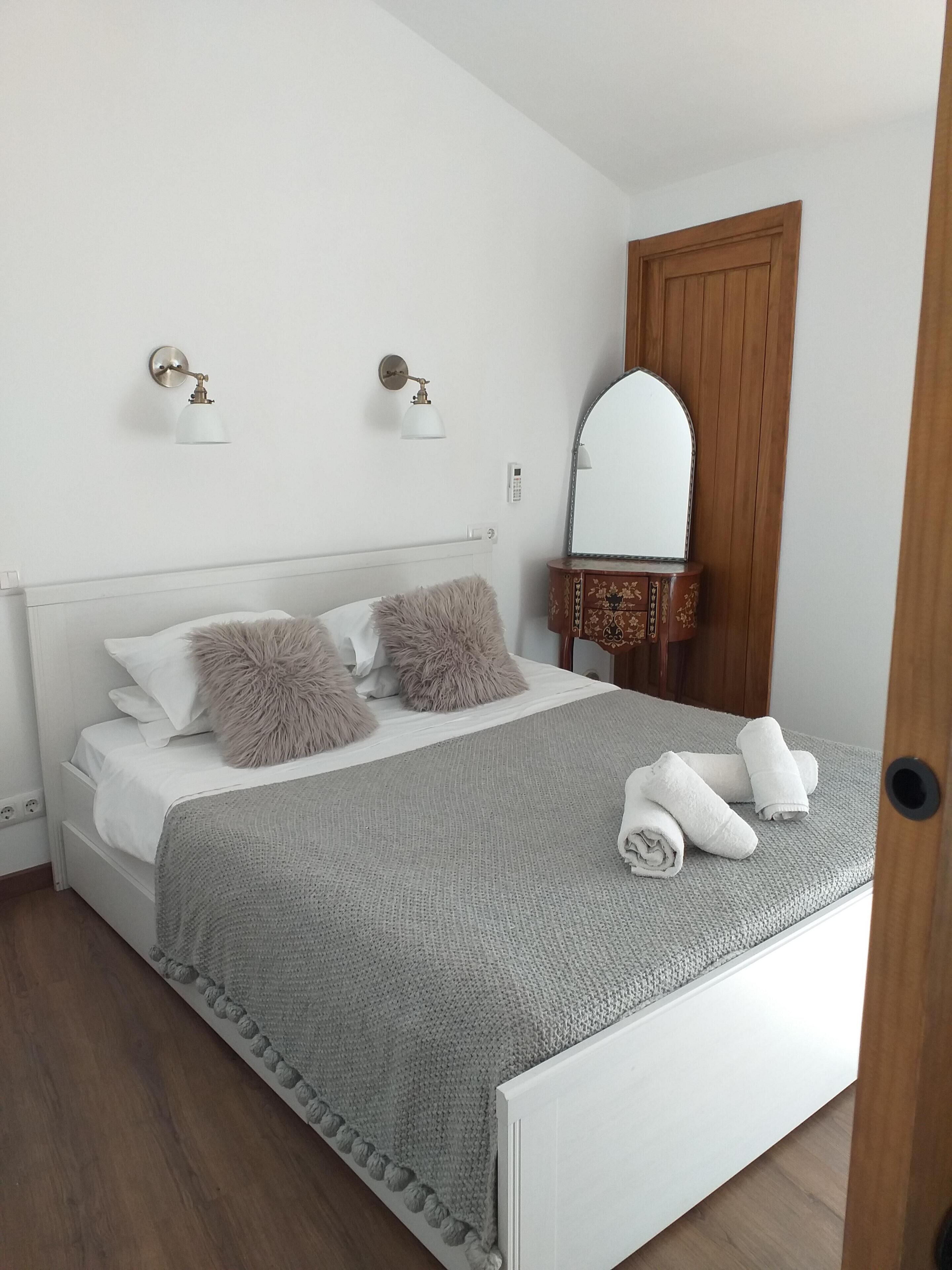 A neatly made bed with fluffy pillows, a grey knitted blanket, and rolled towels on a white headboard.