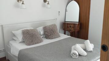 A neatly made bed with fluffy pillows, a grey knitted blanket, and rolled towels on a white headboard.