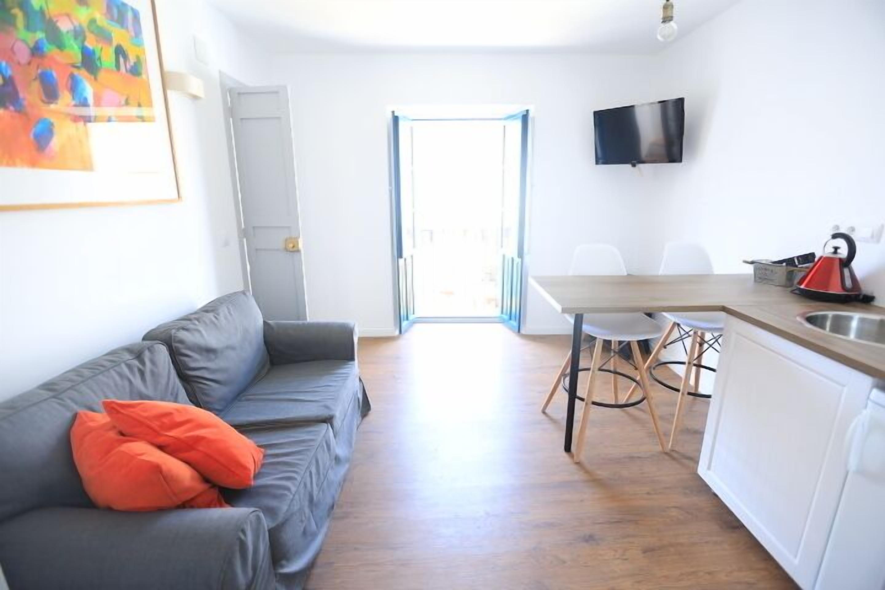 Un apartament modern amb un sofà gris, una cuina amb zona de dinar, i un televisor fixat a la paret.