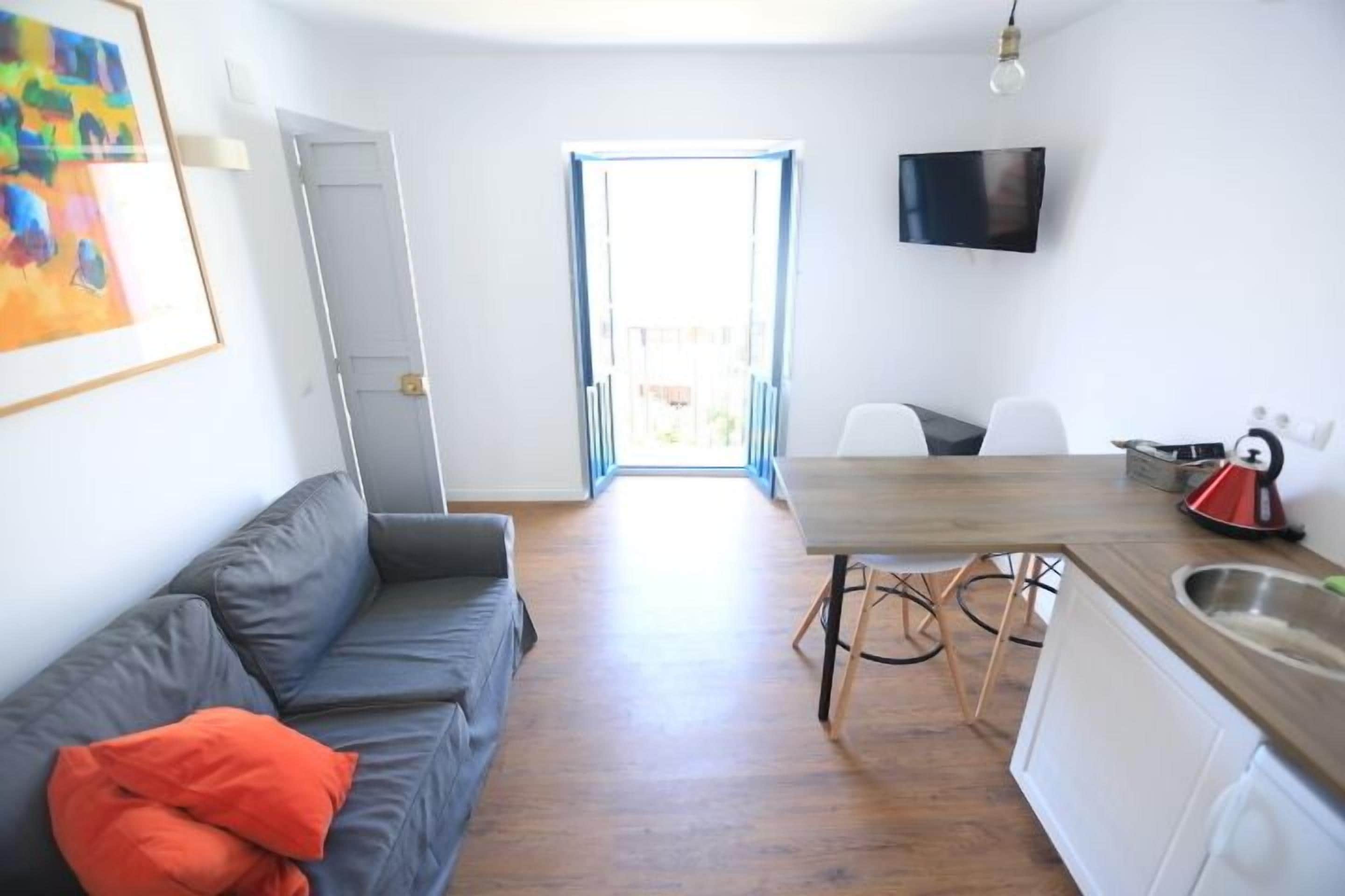Una sala moderna con un sofá gris, una mesa de comedor de madera y una cocina con un lavaplatos y un televisor montado en la pared.