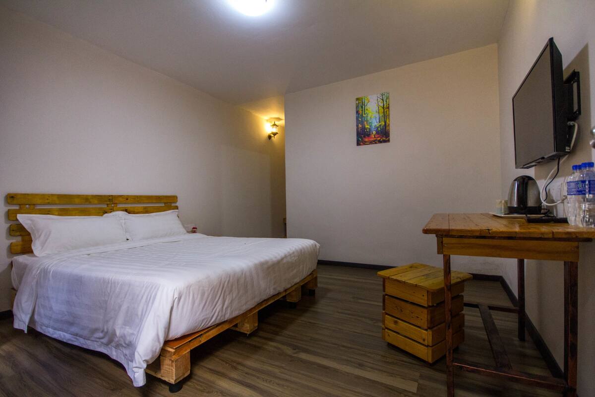 Un dormitorio con una cama de madera, un escritorio con una silla, un televisor y un pequeño cuadro en la pared.
