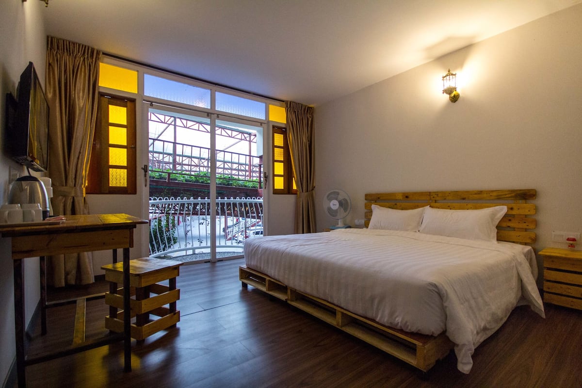 Un dormitorio con cama de madera, un escritorio con silla, un ventilador y vistas a un área exterior con barandilla.