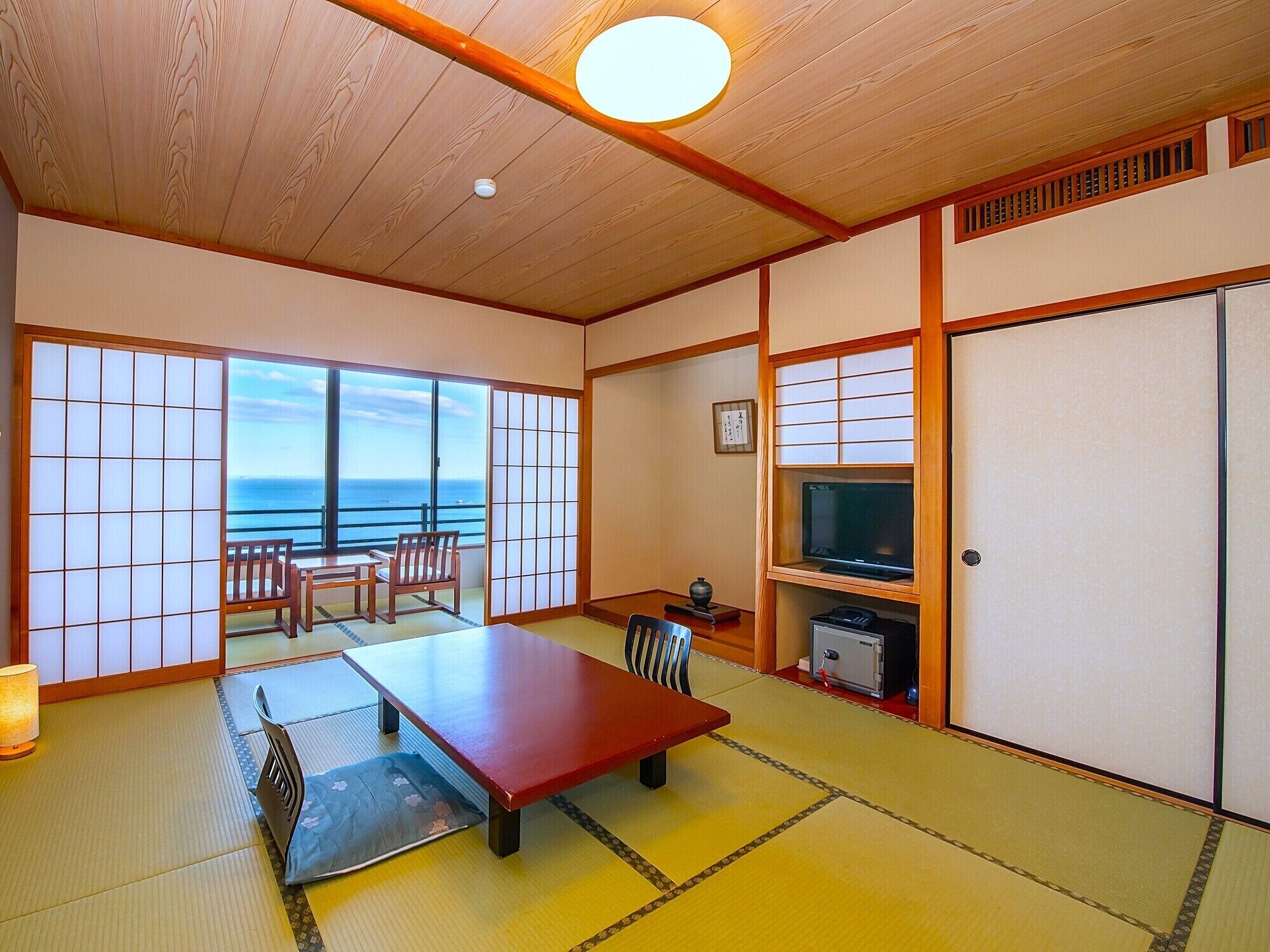 A traditional Japanese room with tatami flooring, a low table, a TV, and a sliding door leading to a balcony with a view of the sea.