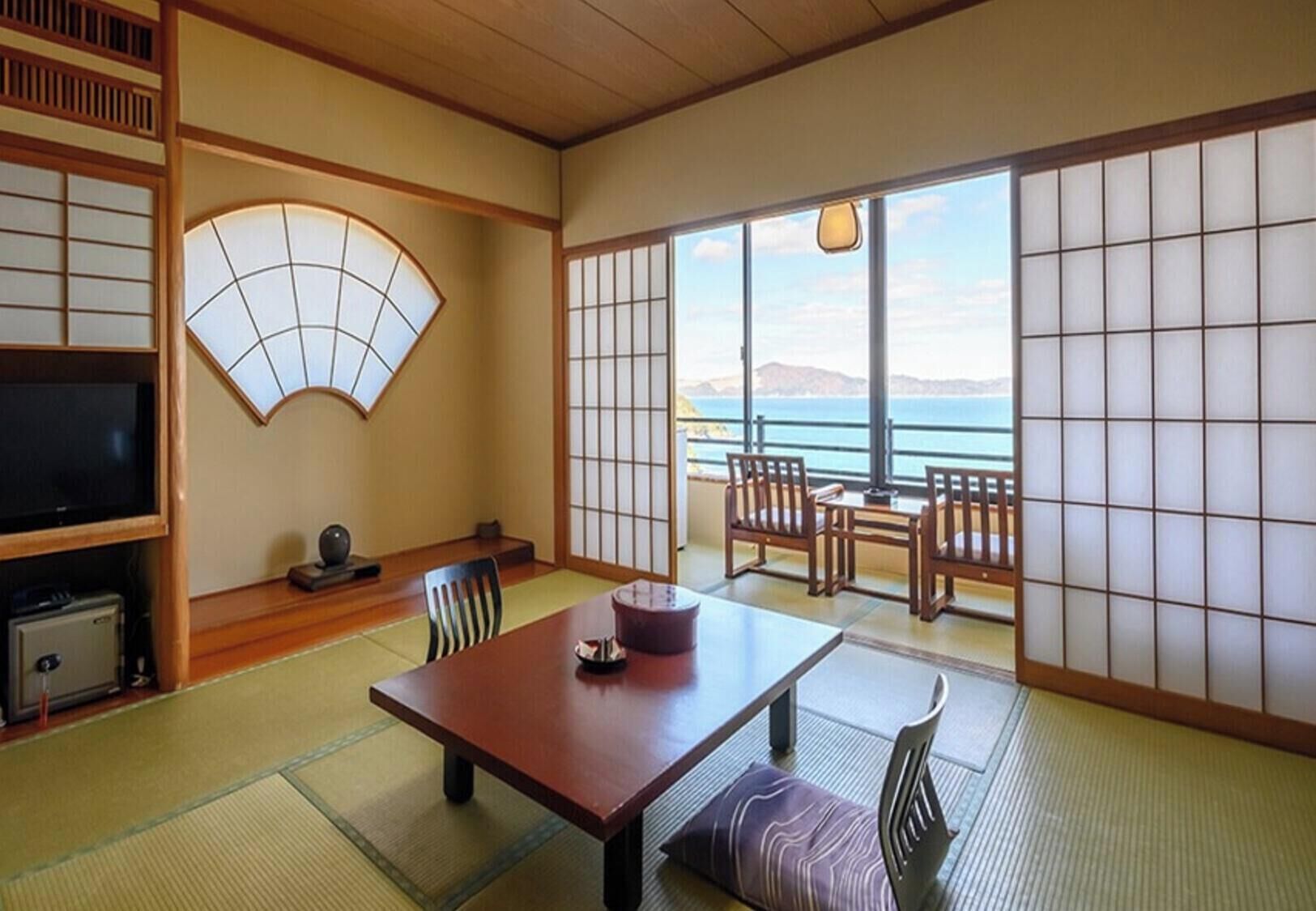 A traditional Japanese room with tatami flooring, a low table with chairs, a television, and a view of the sea.
