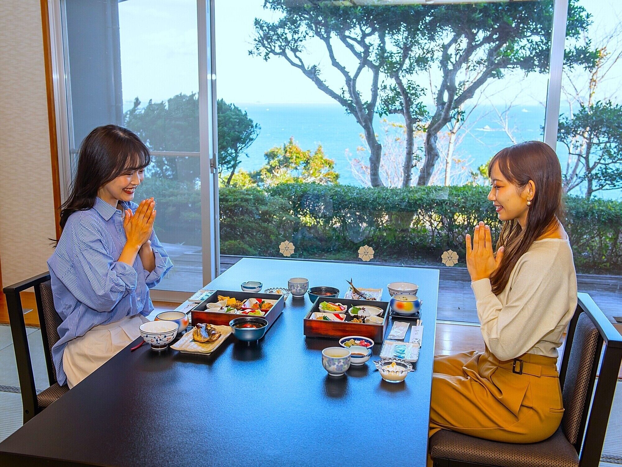 Two people seated at a table with traditional Japanese dishes, a view of trees and the sea through large windows.