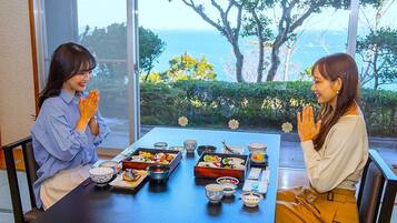 Two people seated at a table with traditional Japanese dishes, a view of trees and the sea through large windows.