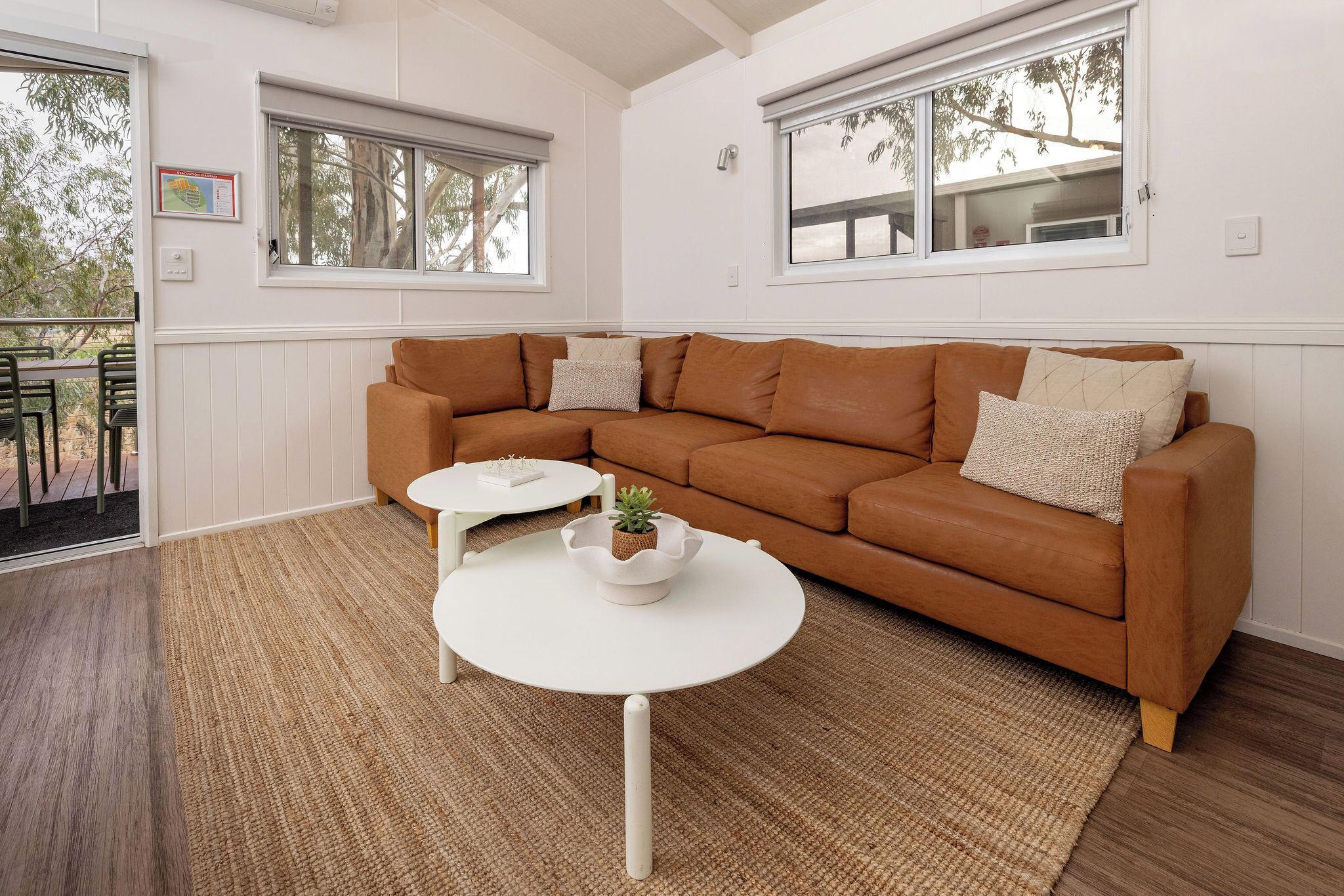A living room with a brown leather sofa, two white side tables, a small white table with a plant, and large windows with blinds.
