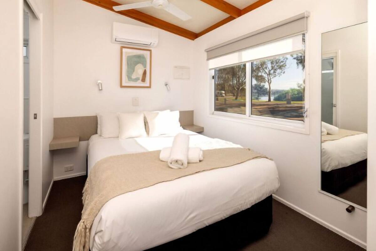 A neatly made bed with a beige blanket and rolled towels, a window with a view of trees, and a ceiling fan.