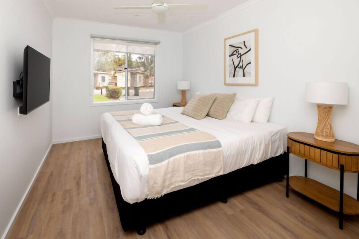 A bedroom with a bed, bedside lamps, a window, and a wall-mounted TV.