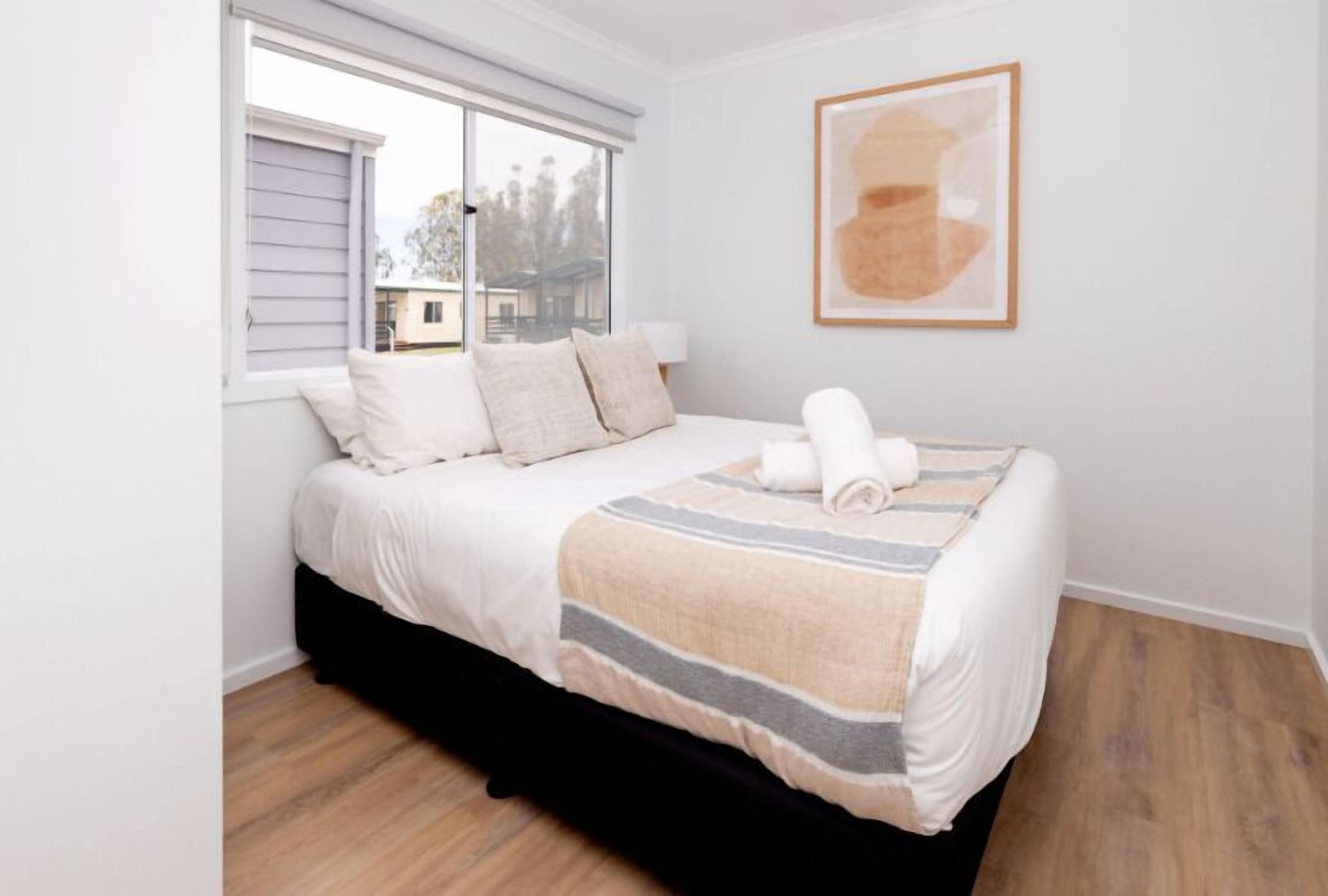 A neatly made bed with a striped blanket, a window with blinds, and a framed abstract painting on the wall.