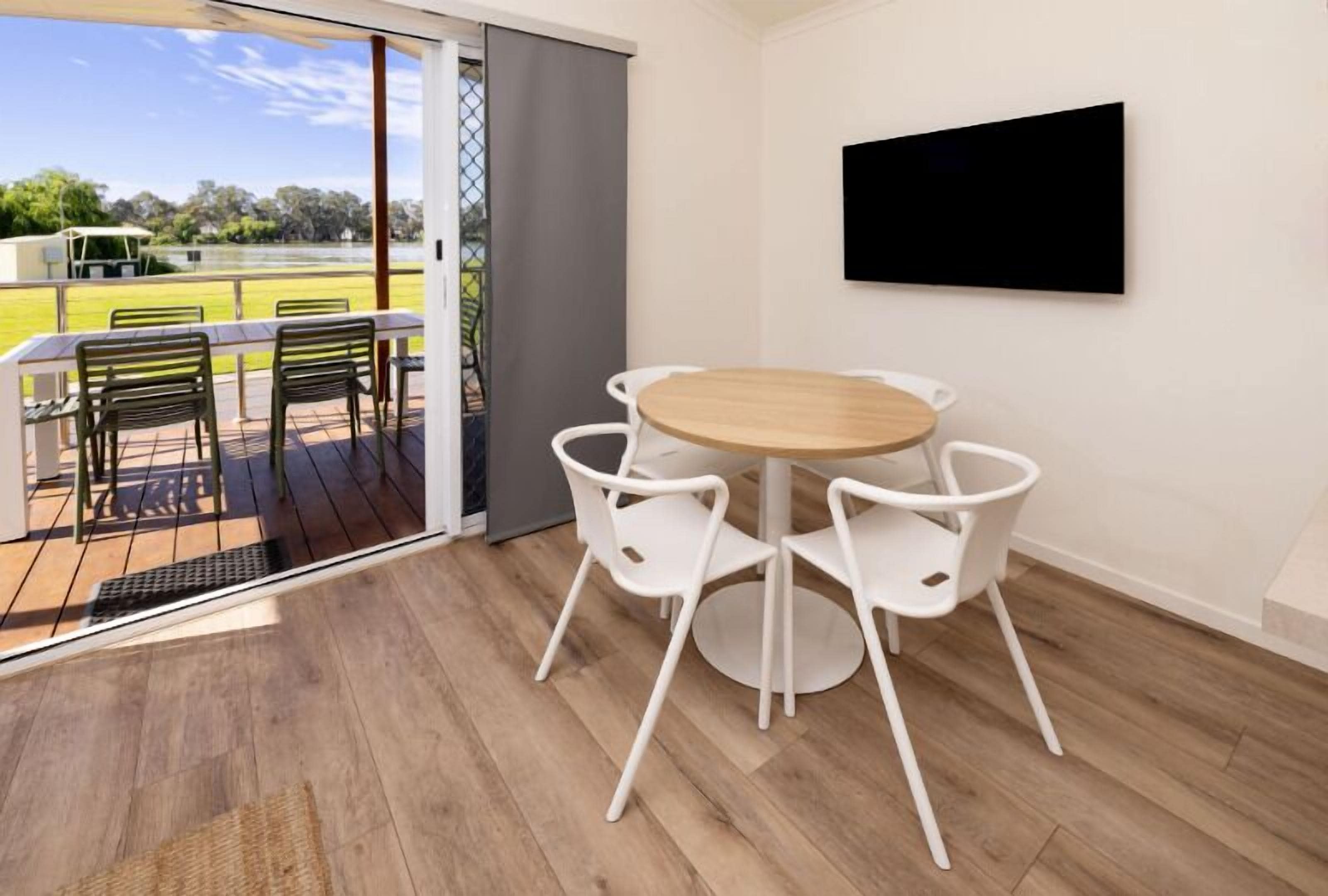 A room with a wooden floor, a round table with white chairs, a TV mounted on the wall, and a view of a grassy area through a glass door.