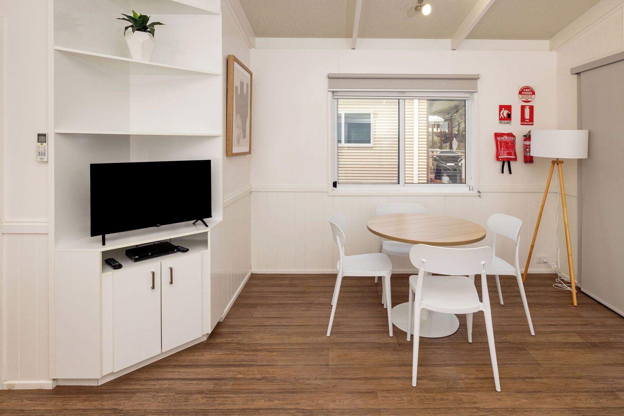 A modern living room with a flat-screen TV, white cabinets, a dining table with chairs, a window with blinds, and a standing lamp.