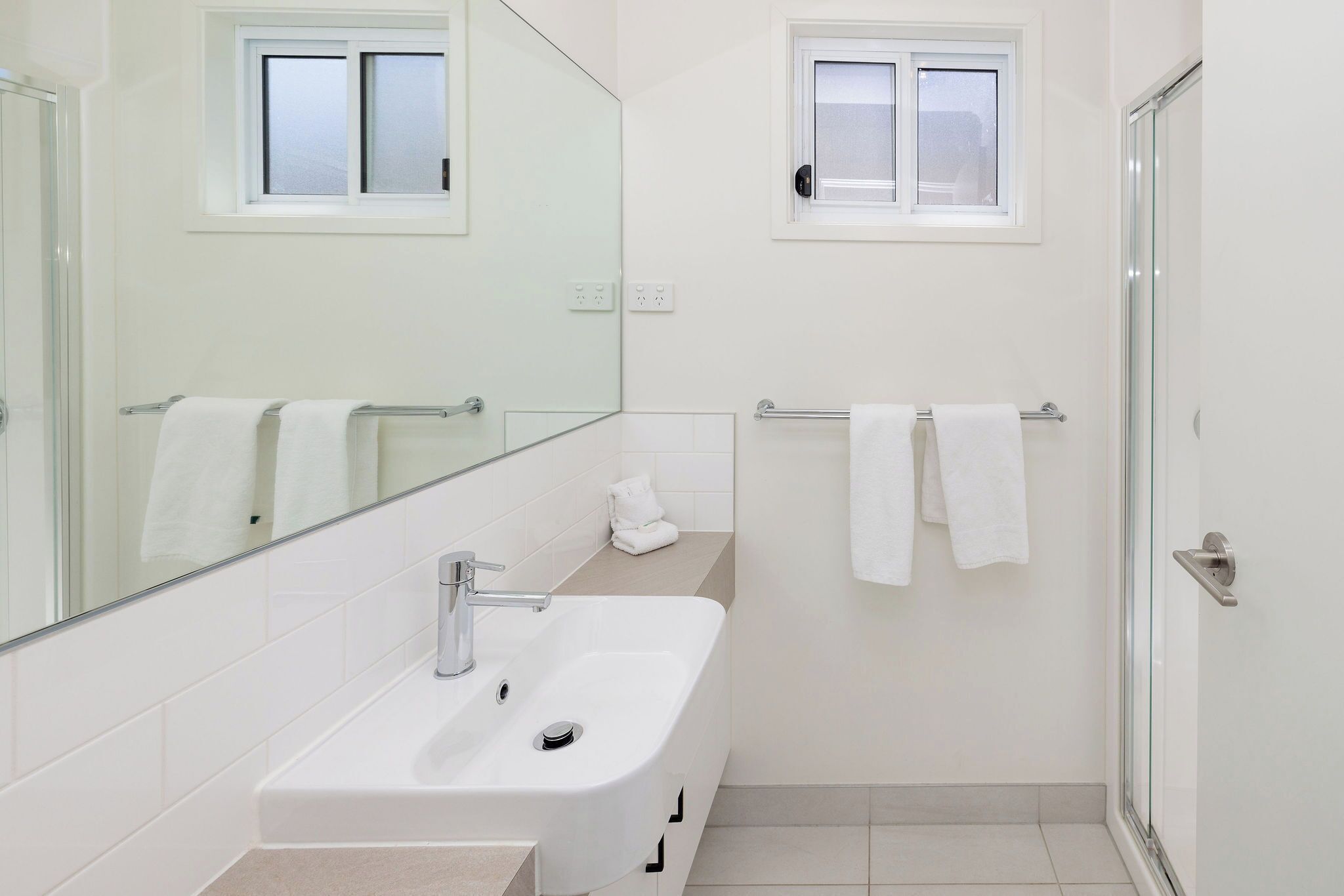 A modern bathroom with a white sink, a glass shower enclosure, and two windows.