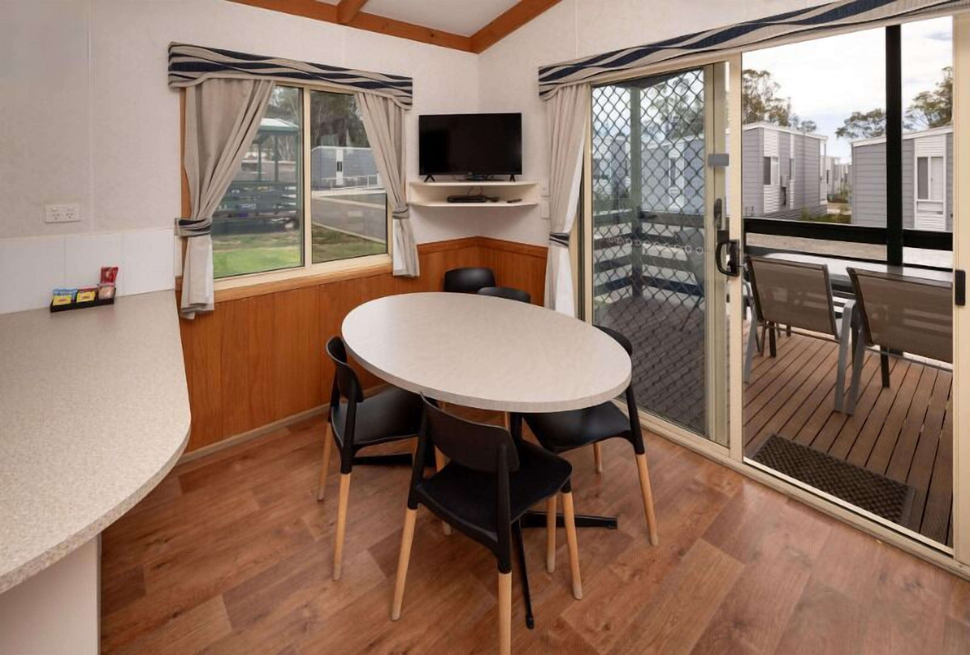 Une pièce avec une table ronde et quatre chaises, un téléviseur posé sur une étagère et une porte-fenêtre coulissante donnant sur une terrasse extérieure.