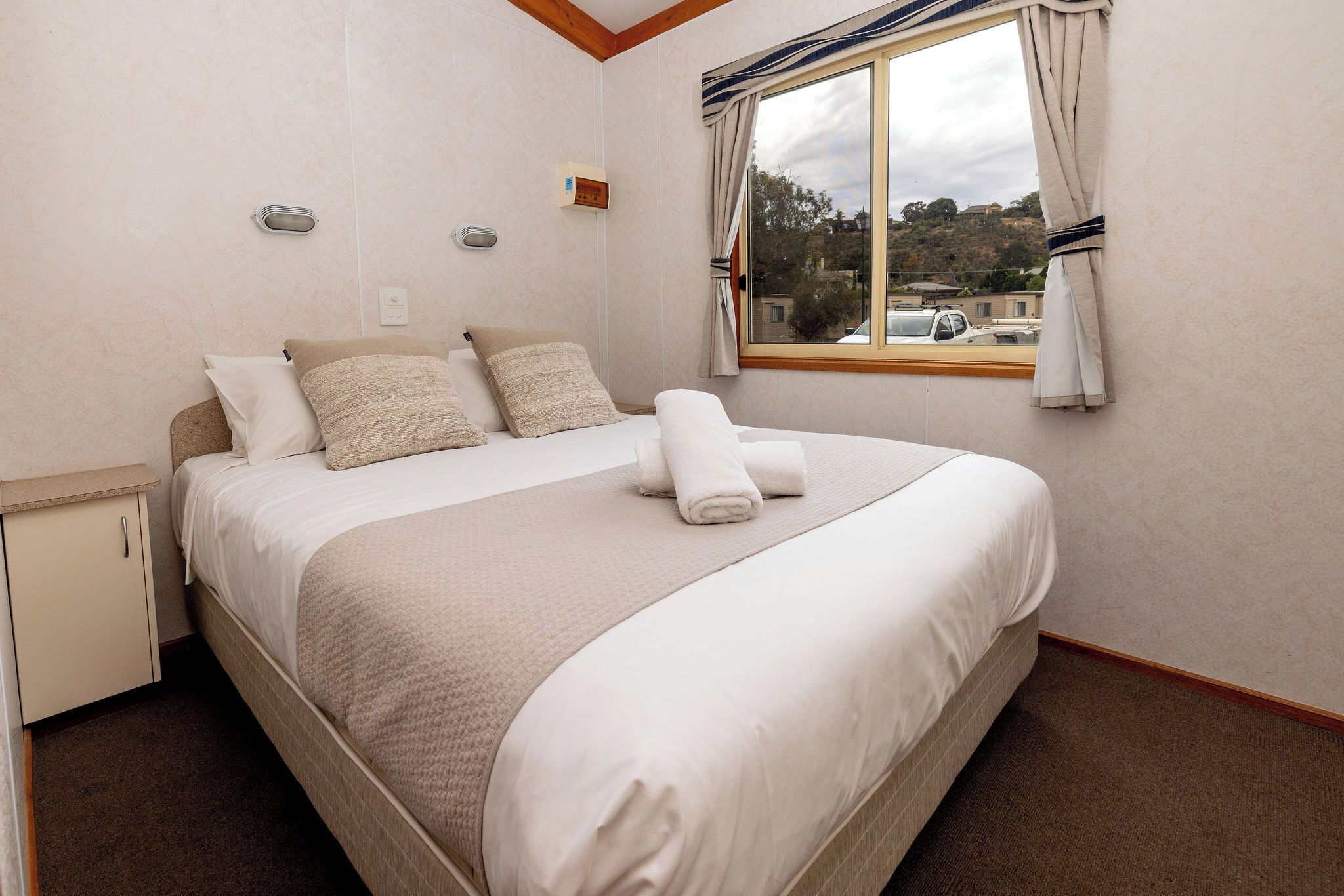 Una cama bien hecha con almohadas y una toalla enrollada, una mesita de noche pequeña, una ventana con cortinas y vistas al exterior.