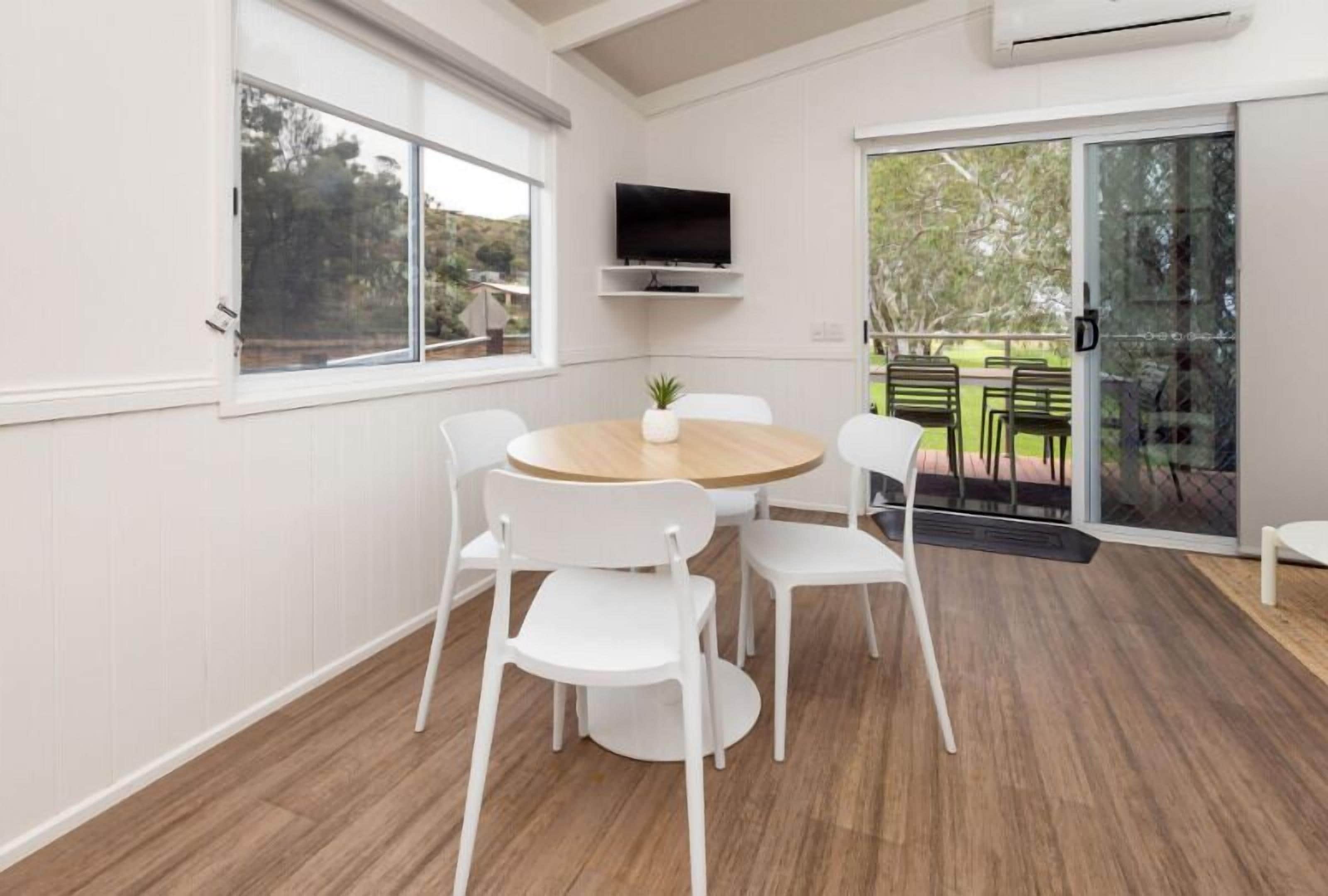 A modern room with a round wooden table, white chairs, a mounted TV, and a sliding glass door leading to an outdoor area.