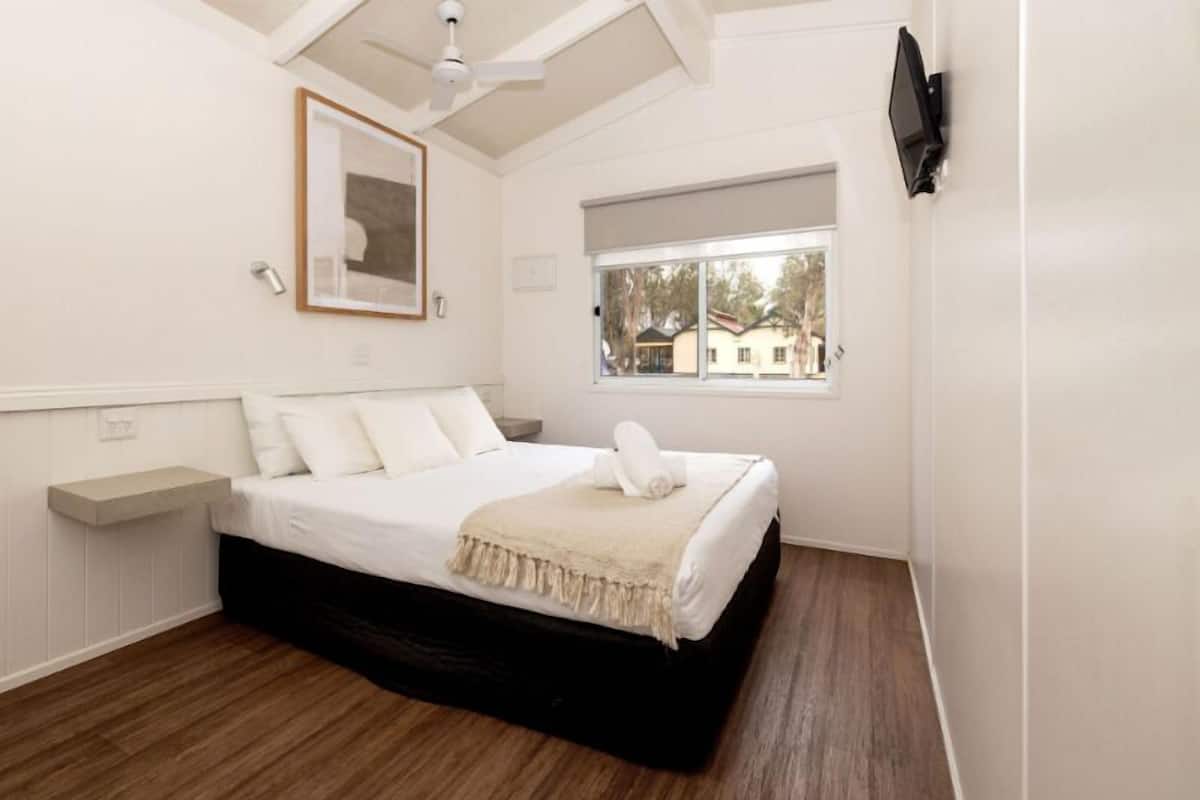 A bedroom with a bed, a window, a ceiling fan, a mounted TV, and a framed picture on the wall.