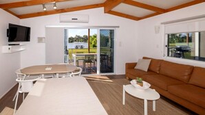 A modern living room with a wooden ceiling, a flat-screen TV mounted on the wall, a dining table with chairs, a brown leather sofa, and a sliding glass door leading to an outdoor patio. - BIG4 Mannum Holiday Park (Mannum)