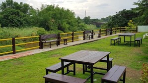 A grassy outdoor area with picnic tables and benches, a yellow railing, and a pathway. - Baansri Family (Kabin Buri)