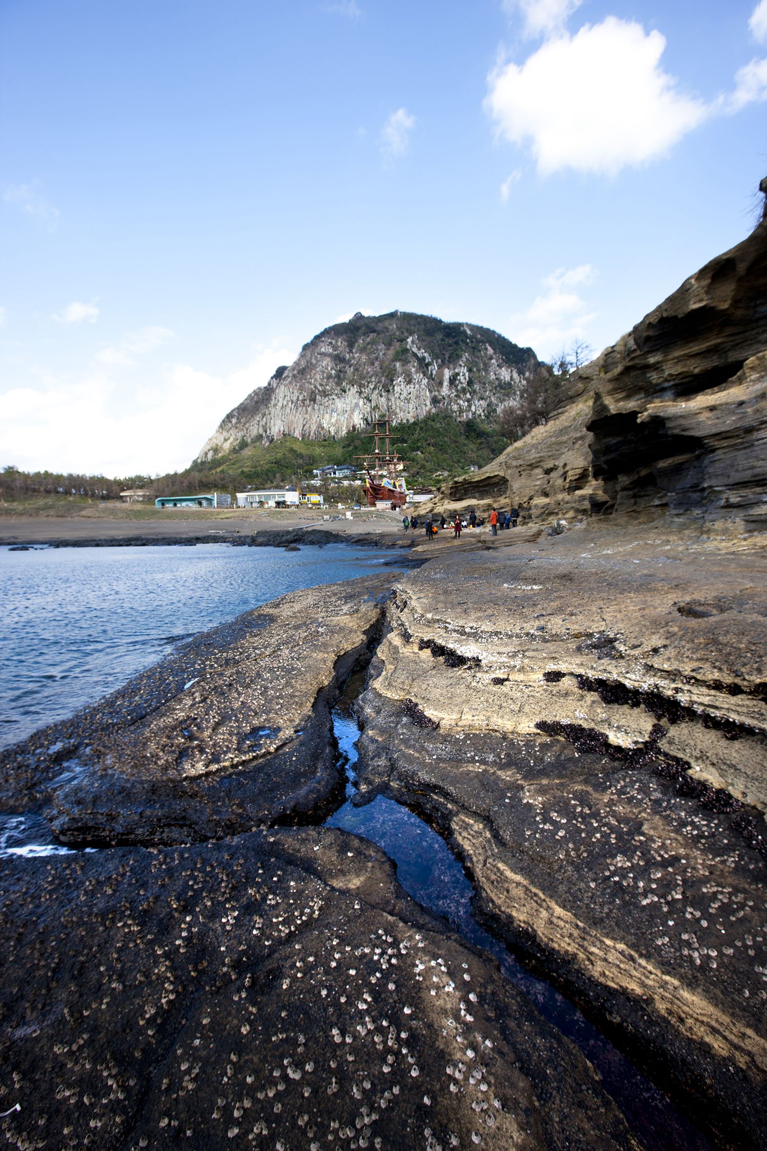 A rocky shoreline with a mountain in the background and a few buildings near the water.