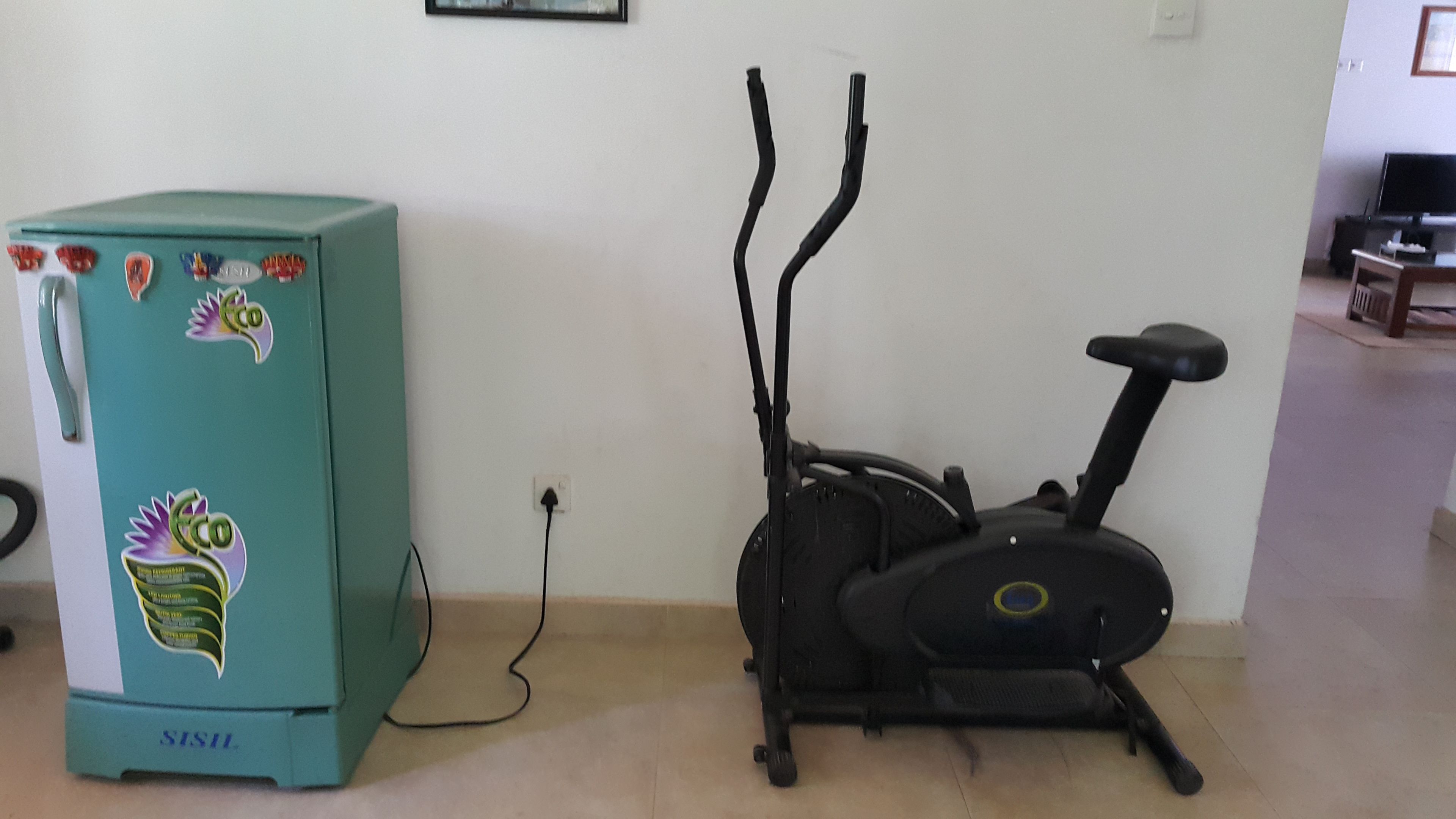 Ein türkis-blauer Kühlschrank mit Aufklebern, ein Heimtrainer und eine Steckdose an der Wand.