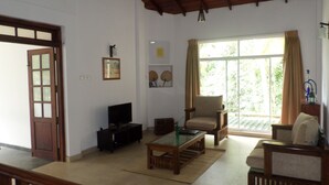A living room with a television, a sofa, a chair, and a coffee table. There is a window with curtains, a door, and a ceiling fan.