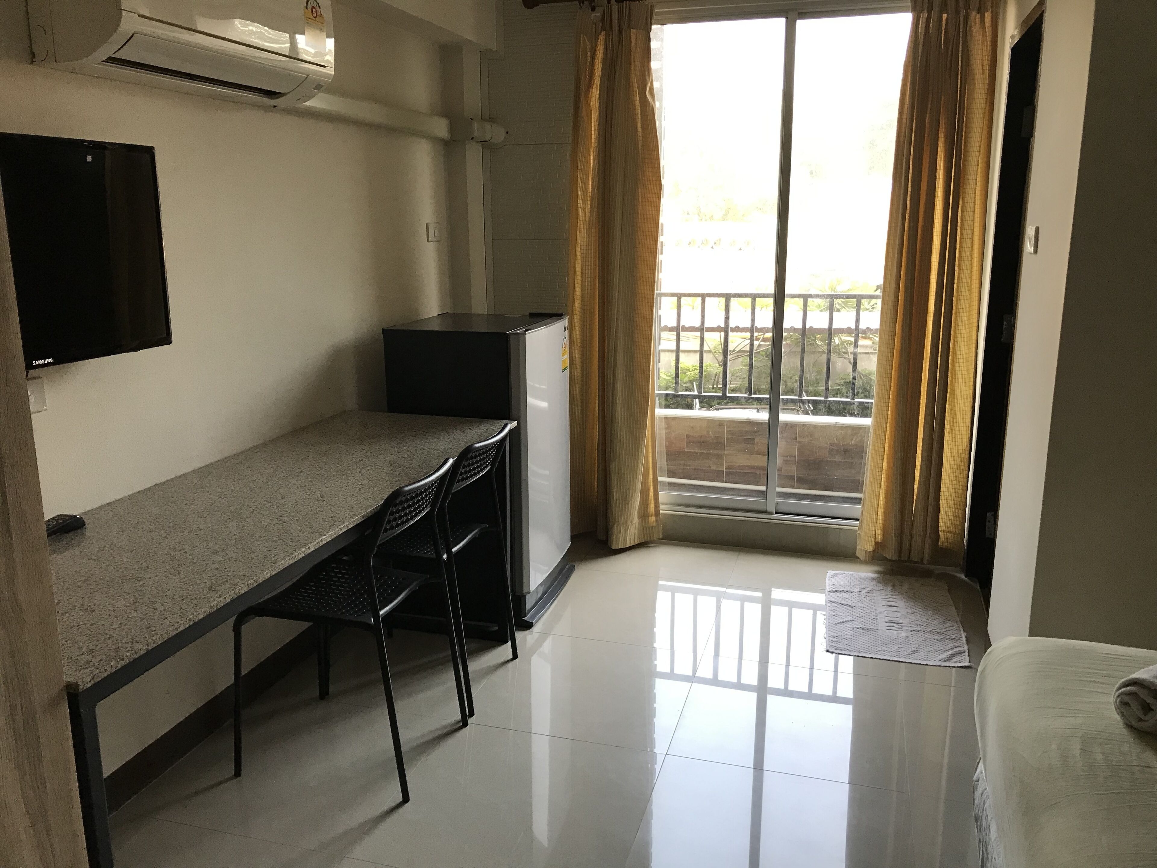 A small, clean room with a kitchenette, a flat-screen TV, a desk with two chairs, and a sliding glass door leading to a balcony.