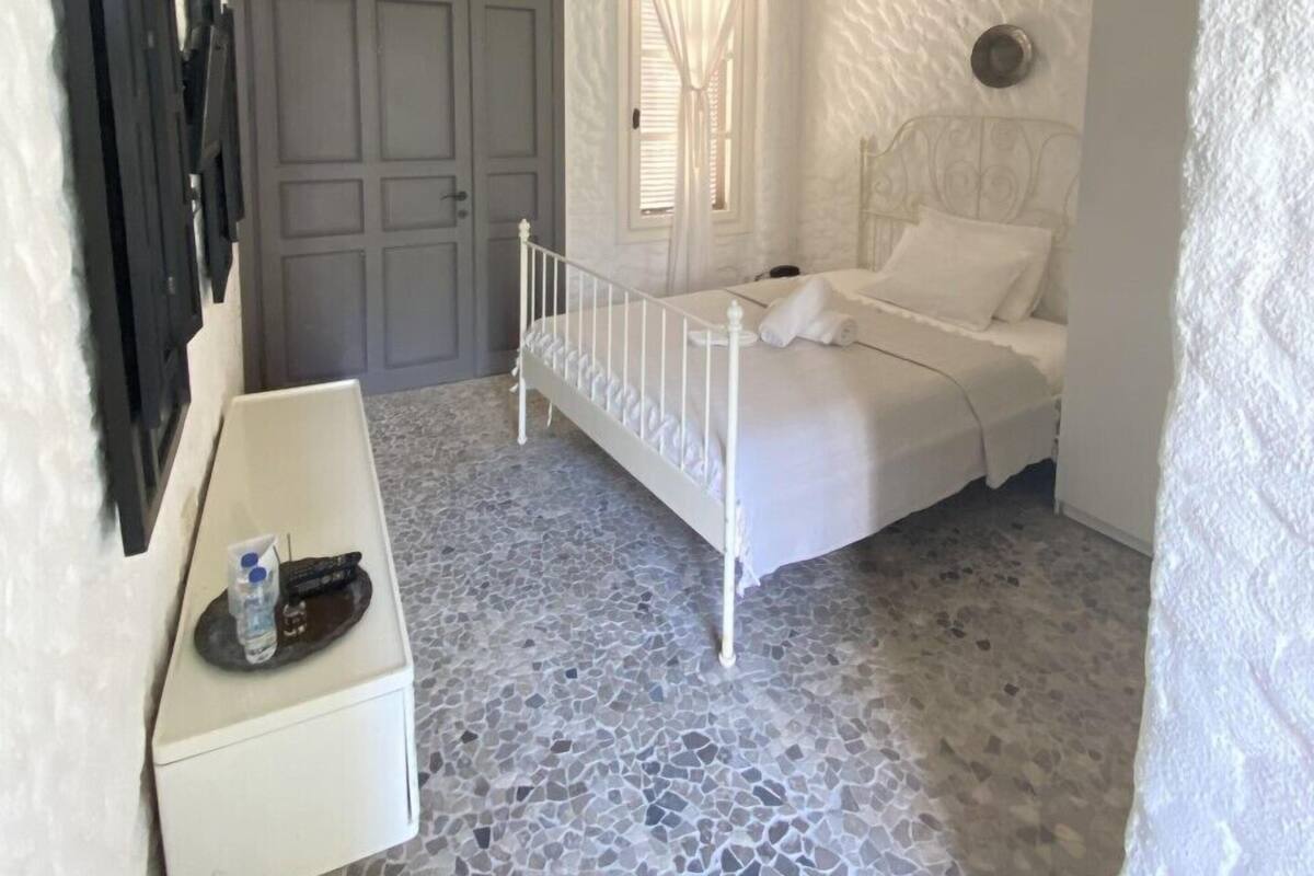 A bedroom with a white bed, a ceiling fan, a door, and a small white table with a bottle and remote.