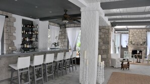 Zona de bar con pared de piedra, taburetes blancos y un ventilador de techo.