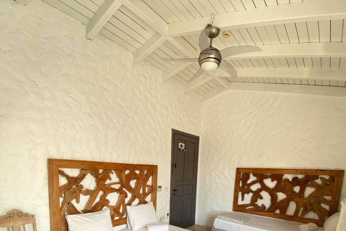 A room with two beds, a wooden headboard, a small wooden chair, a ceiling fan, and a stone-tiled floor.
