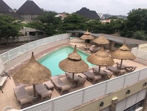 Un área de piscina en la azotea con sillones de descanso y sombrillas de paja.