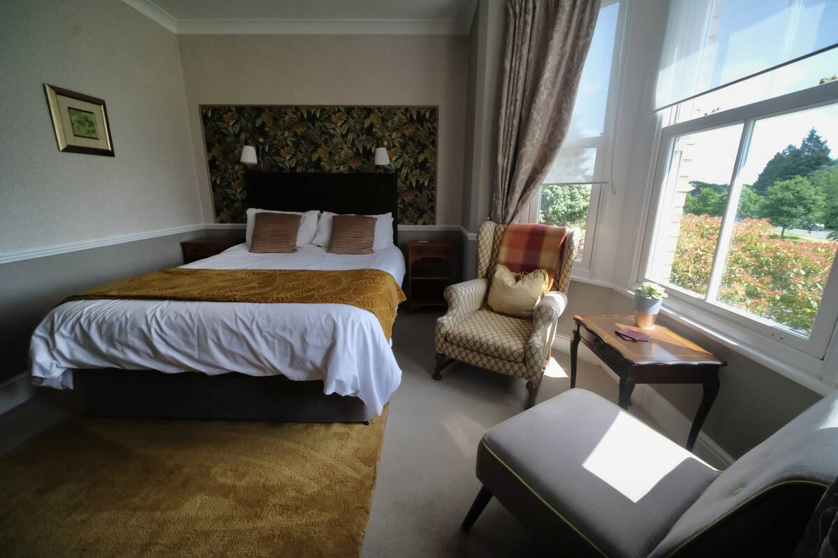 Habitación de hotel con una cama grande, una silla, una mesita y una ventana con vistas a zonas verdes.