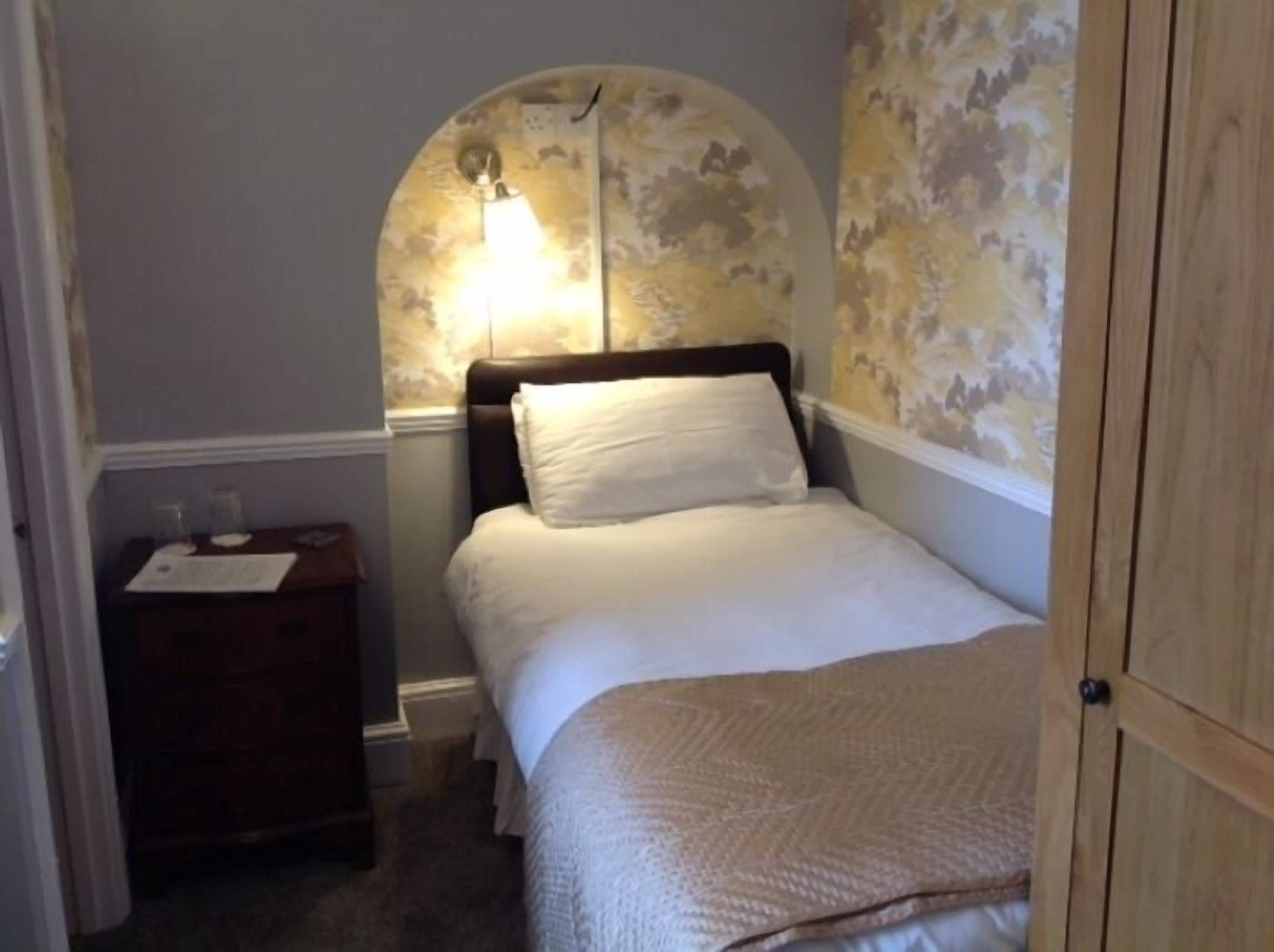 A small, cozy bedroom with a single bed, a nightstand, and a wall-mounted lamp.