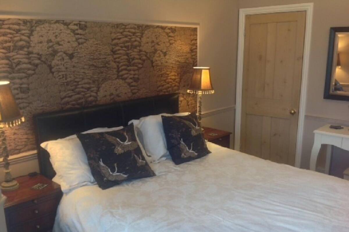 Una cama bien hecha con ropa de cama blanca y dos almohadas decorativas, un cabecero de textura de piedra, dos lámparas de noche y una puerta de madera.