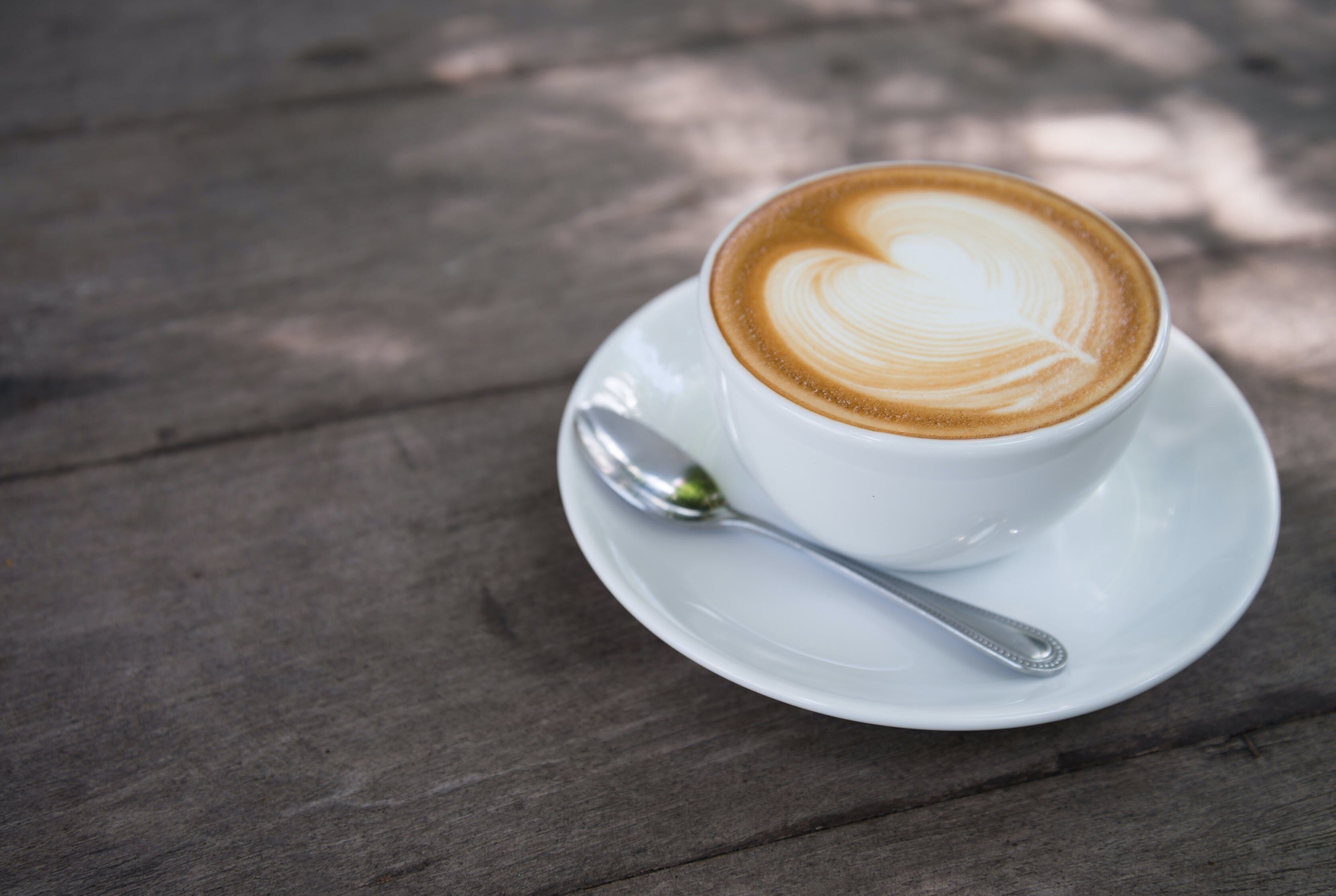 A white cup of coffee with latte art on a white saucer and spoon, placed on a wooden table.