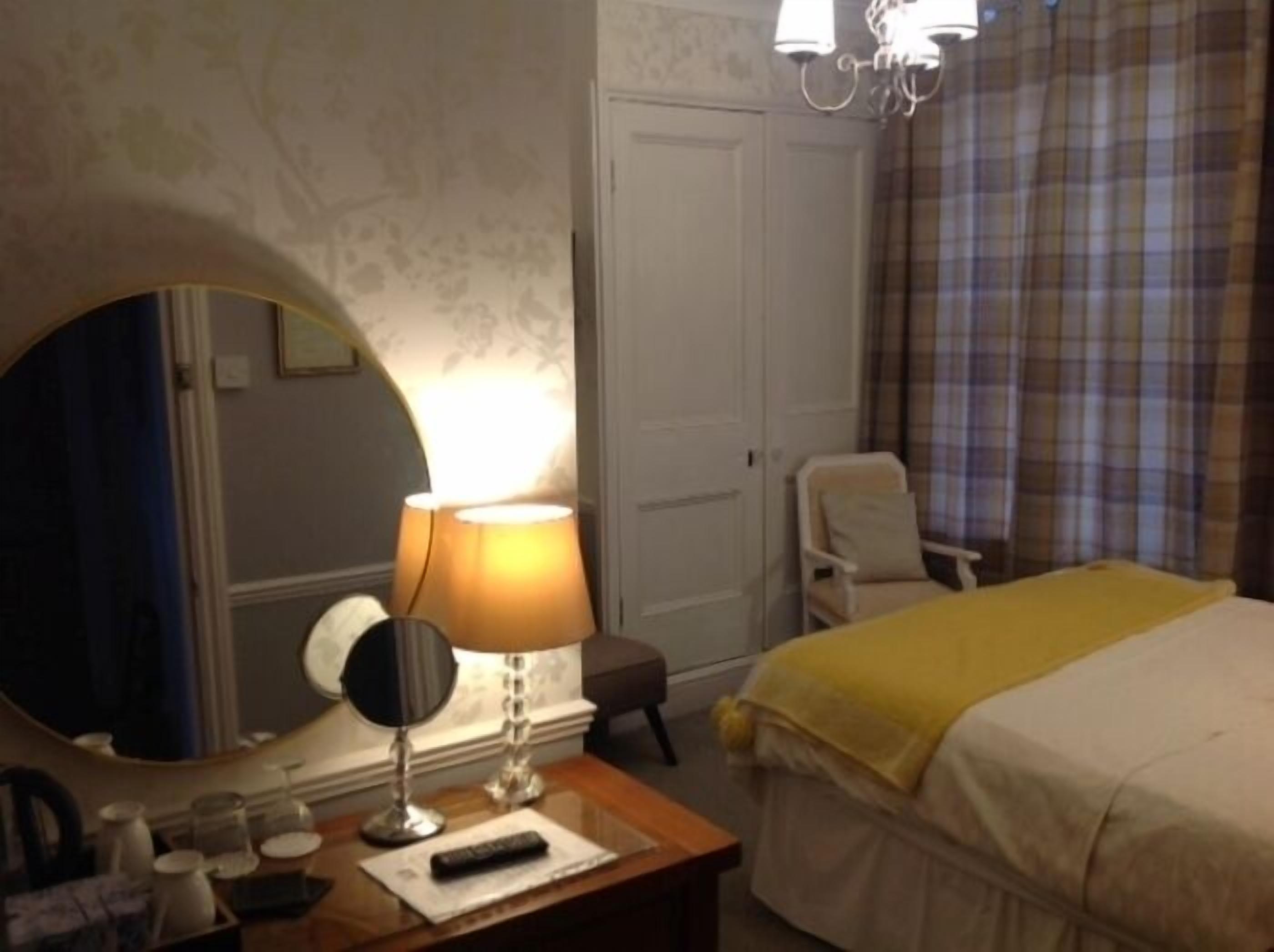 A hotel room with a bed, a bedside table with a lamp, a mirror, a chair, and a door.