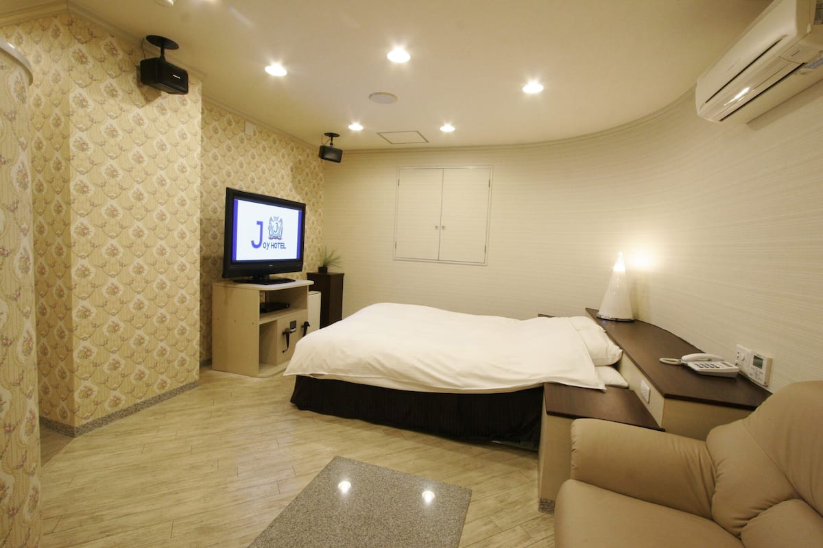 A hotel room with a bed, a desk with a lamp, a television, and an air conditioning unit.