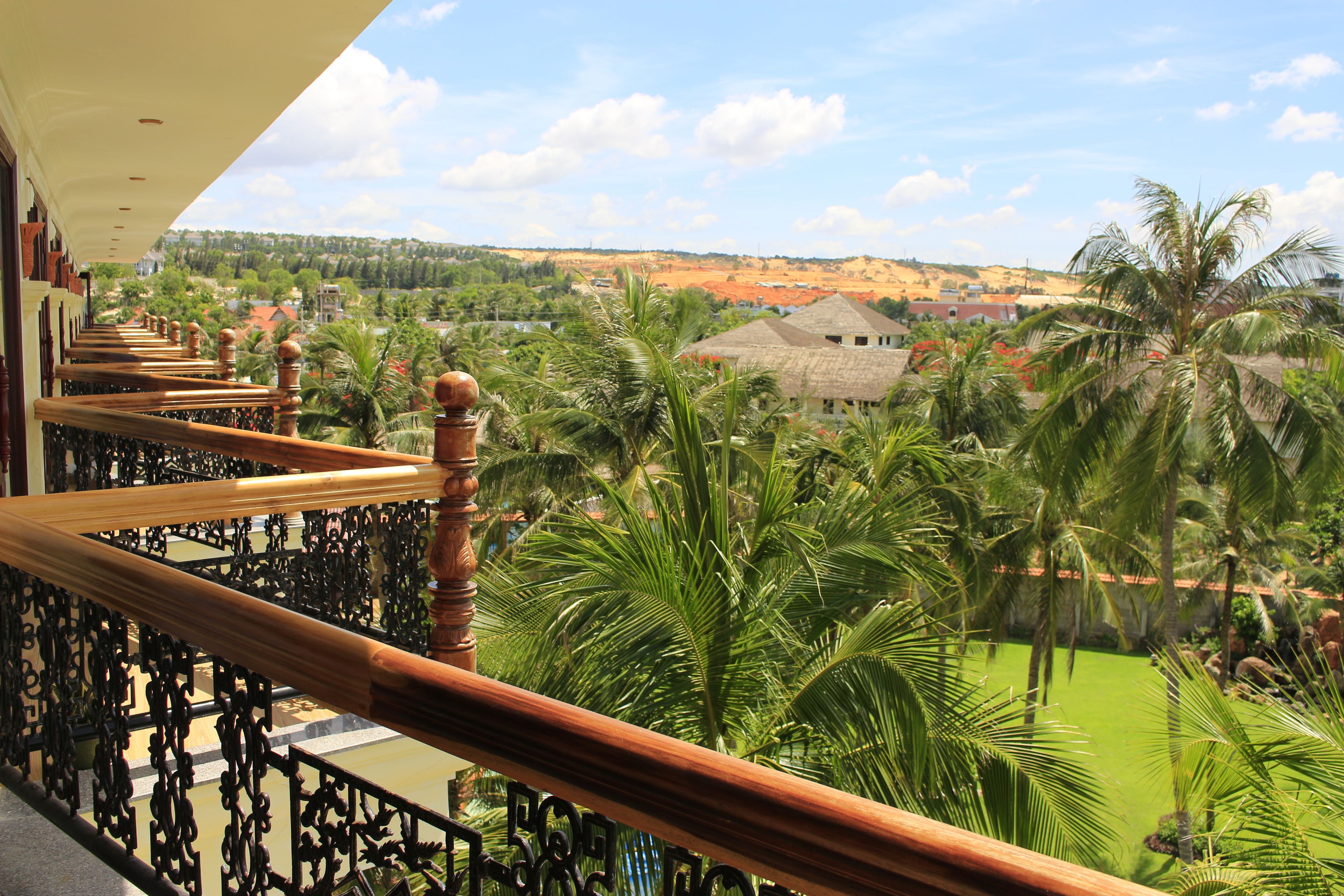 A balcony with a wooden railing overlooking a lush garden with palm trees and a view of a town in the distance.