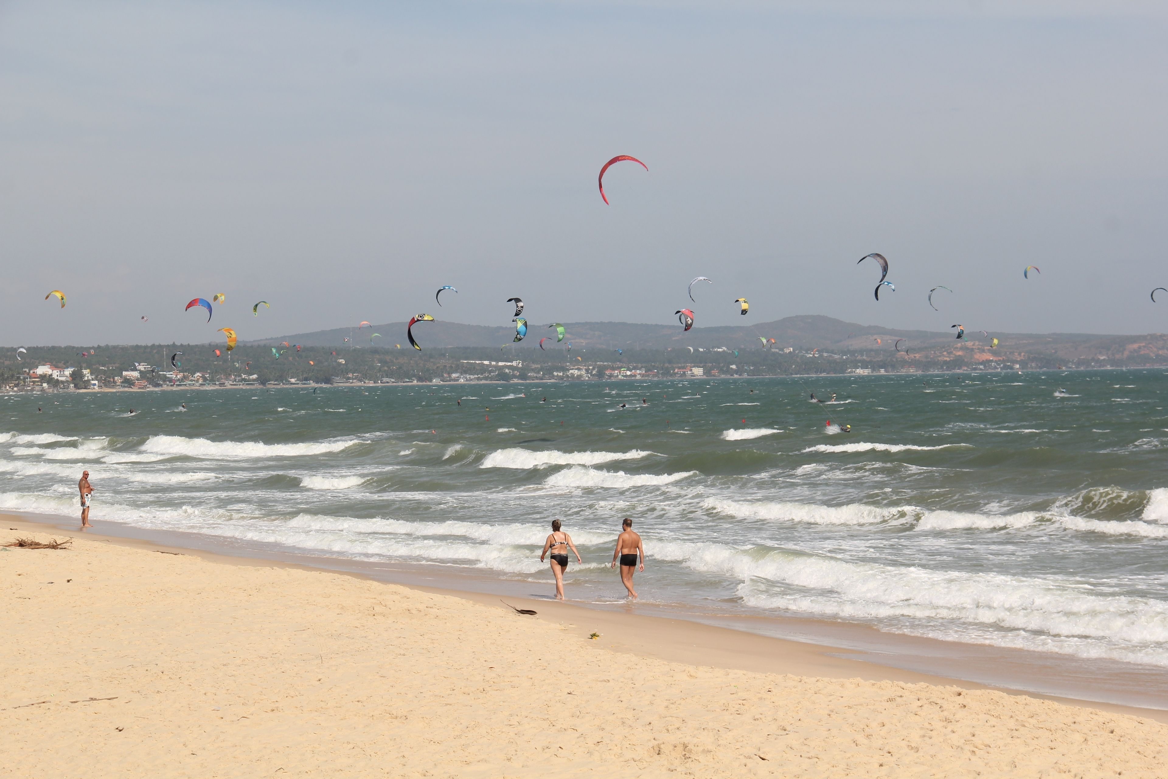 Uma praia com pessoas caminhando pela margem e pipas voando no céu.