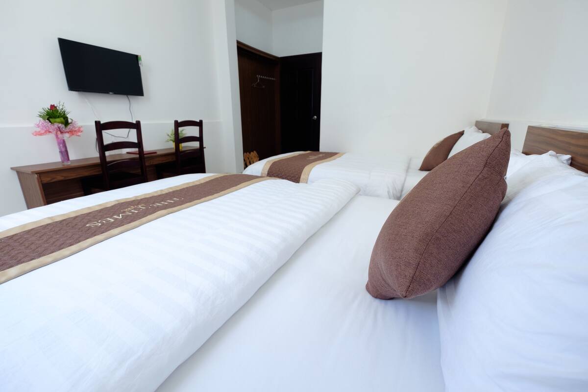Habitación de hotel con dos camas, un televisor montado en la pared, un escritorio con sillas y una puerta que da a otra habitación.
