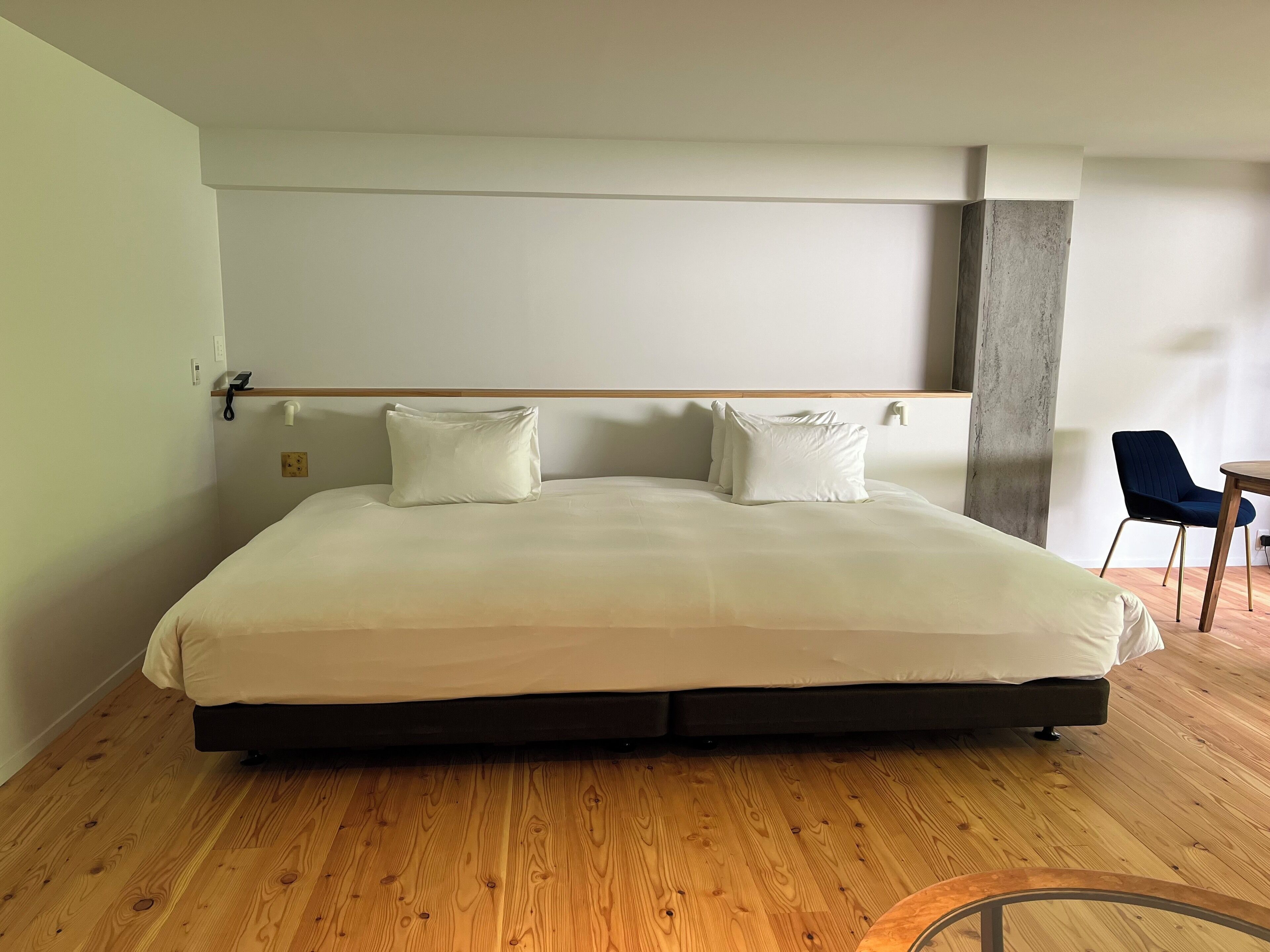 A large bed with white linens, a wooden floor, and a small table with a chair in the corner.
