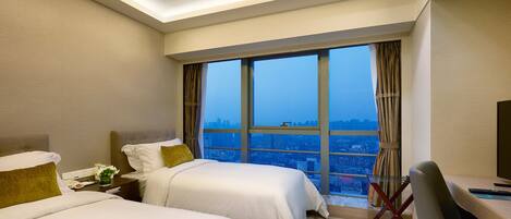 A hotel room with two beds, a desk, and a large window offering a city view.