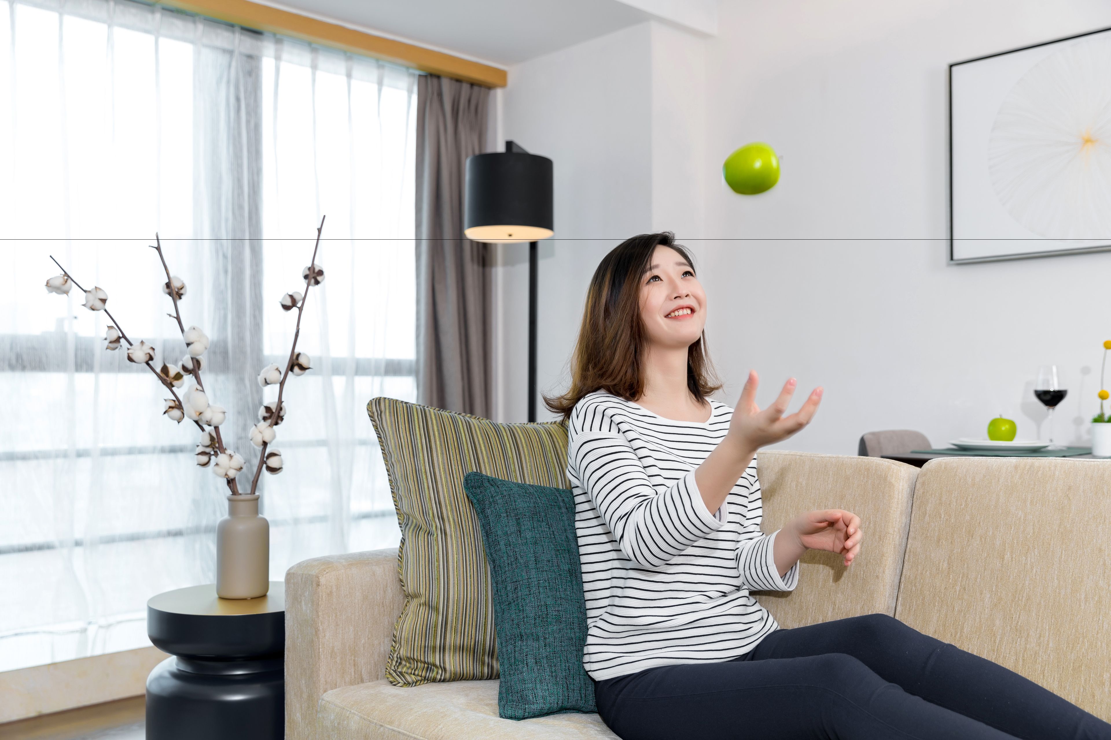 Een vrouw zit op een bank in een moderne woonkamer, met een vaas vol takken, een lamp en een ingelijst beeld aan de muur.