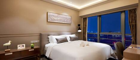 Quarto de hotel moderno com uma cama grande, uma escrivaninha e vista para a cidade à noite.